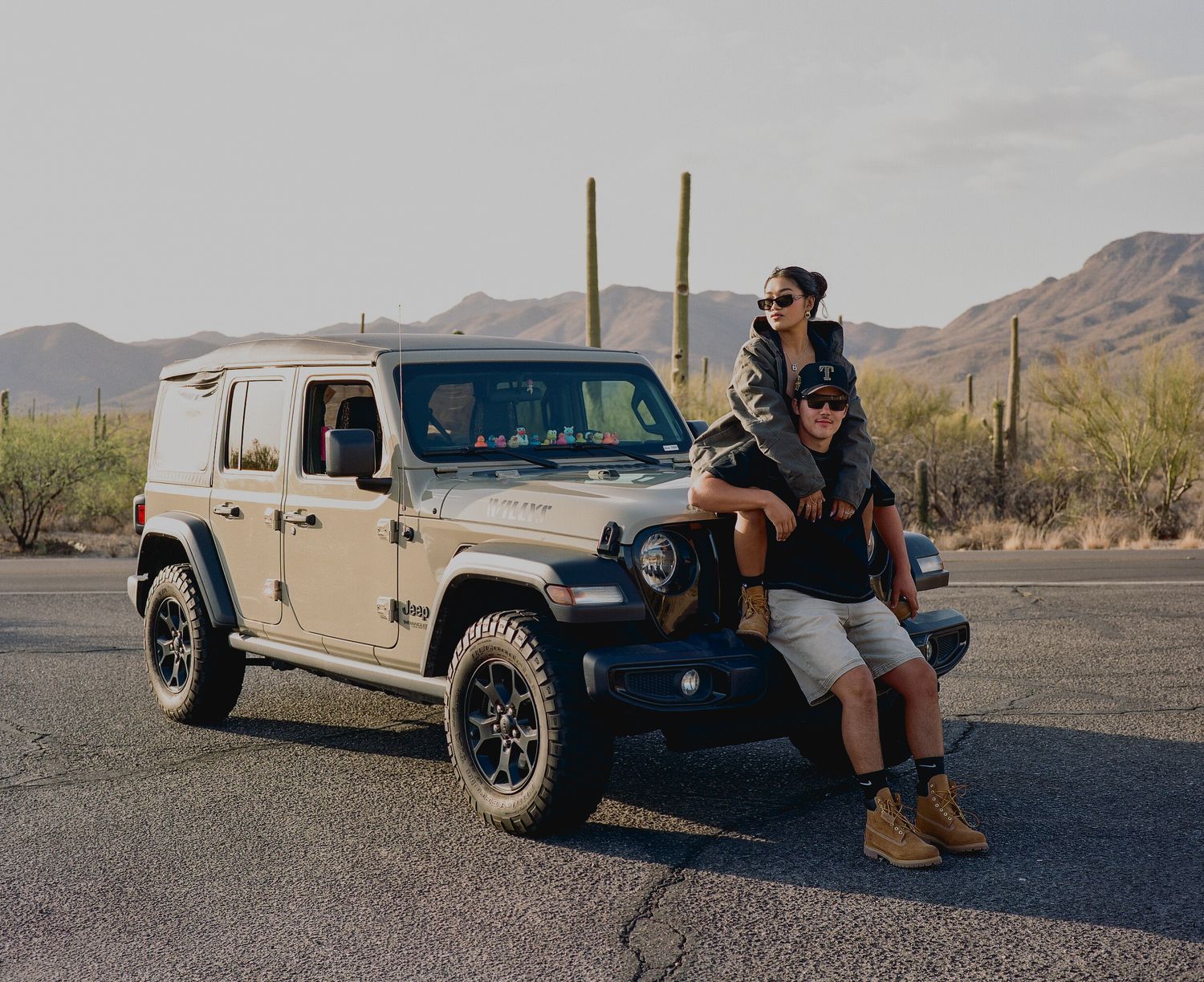 Couple posing together leaning on beige Jeep Wrangler on desert road with cacti backdrop.