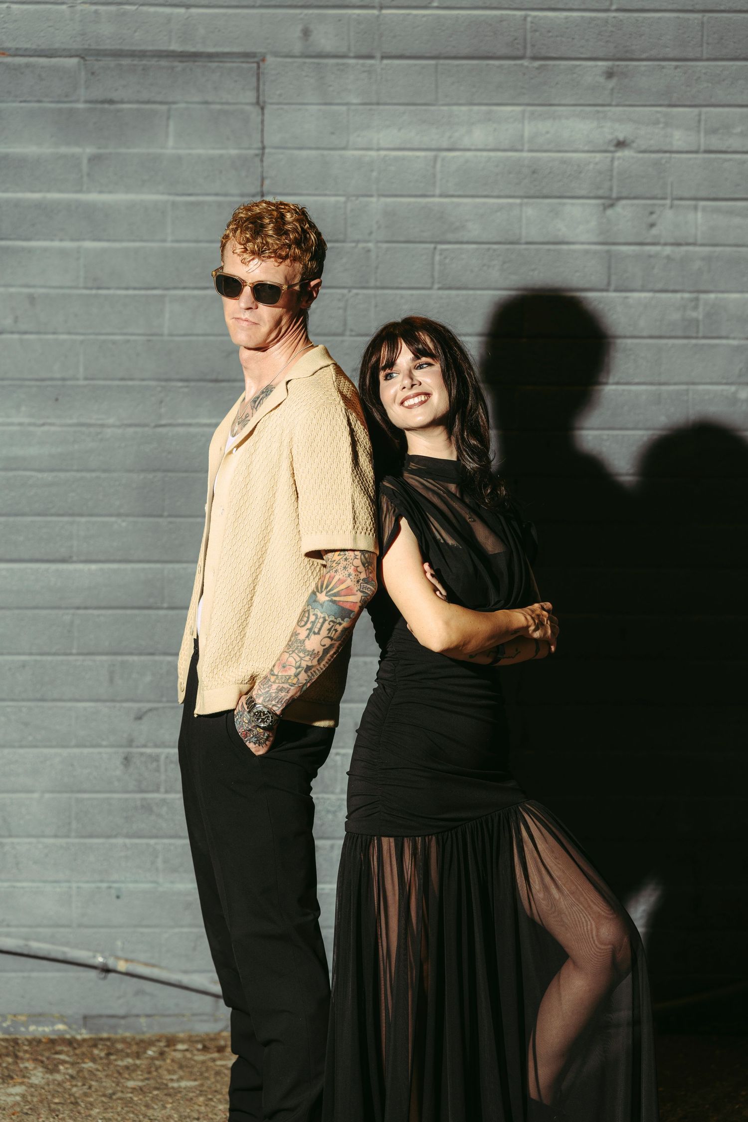 Stylish couple posing against gray brick wall, man in sunglasses with tattoos, woman in black sheer dress.