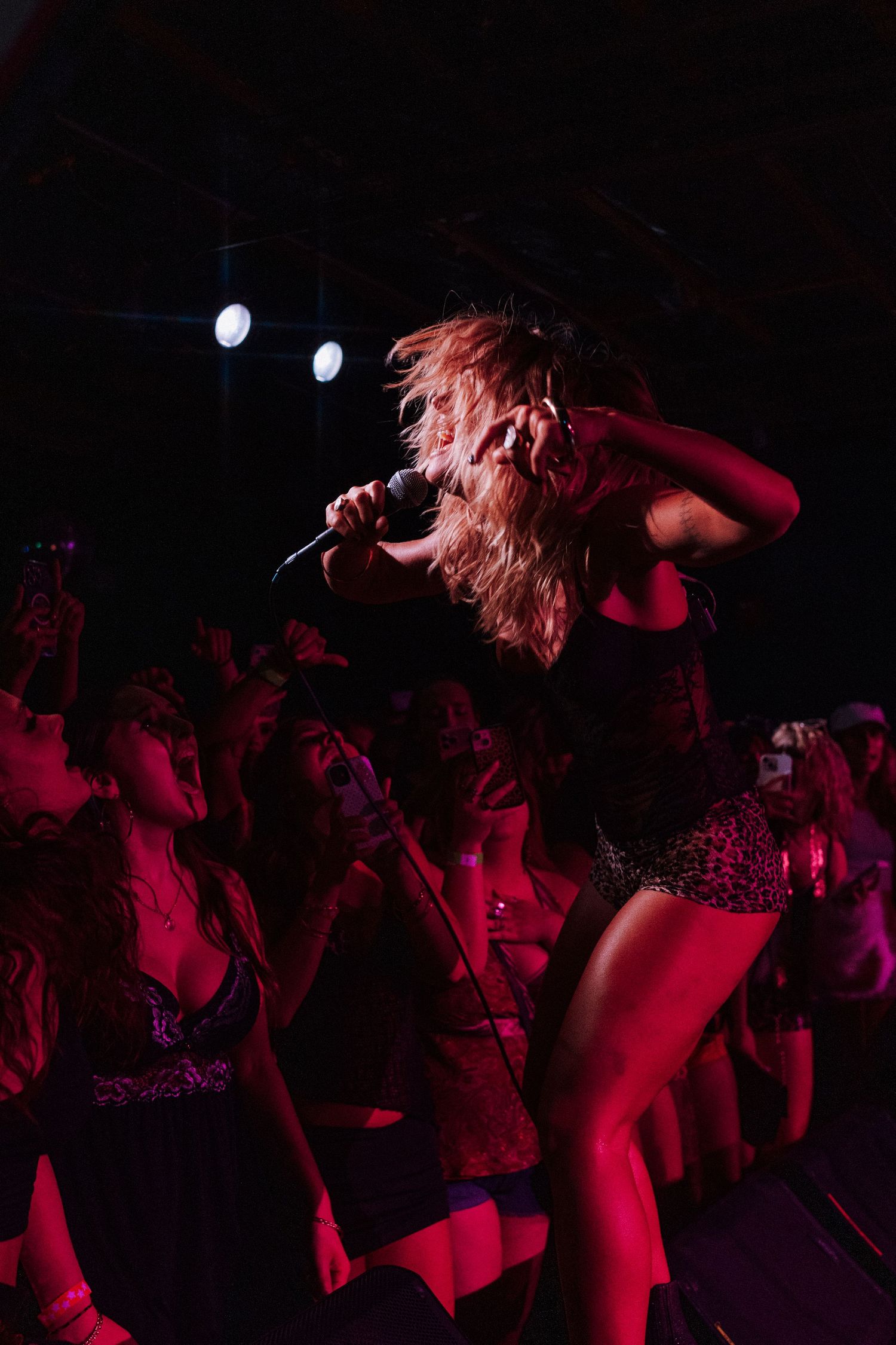 A female performer sings into a microphone on stage, hair flying, bathed in red light with an energetic crowd behind her.