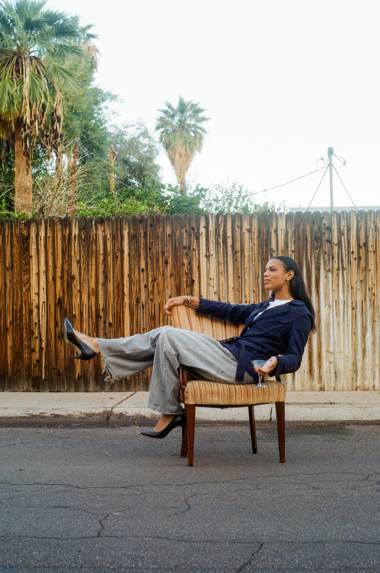 Woman in navy blazer and gray trousers relaxes on chair outdoors near wooden fence.