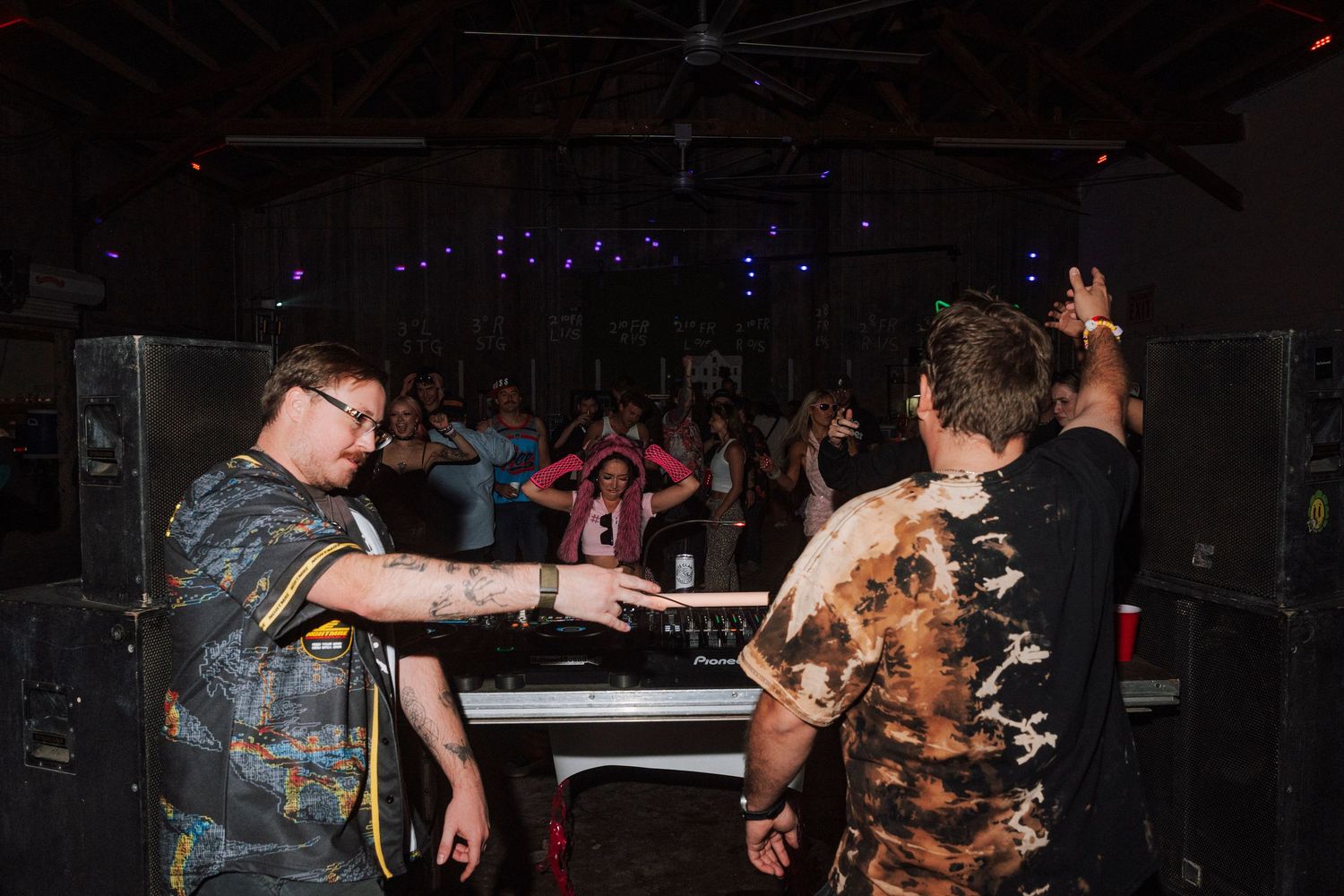 Two DJs performing at a mixing board for a large crowd in a dimly lit event venue.