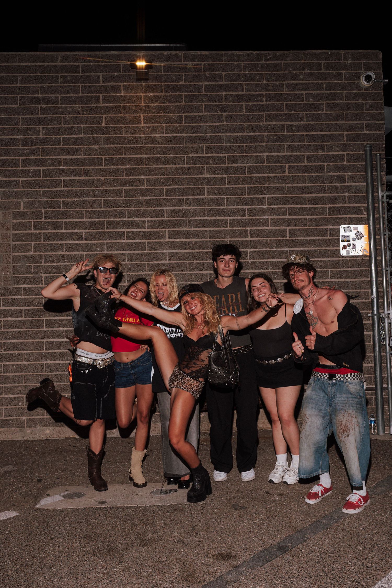 Group of young friends posing energetically against a brick wall at night in edgy festival outfits.