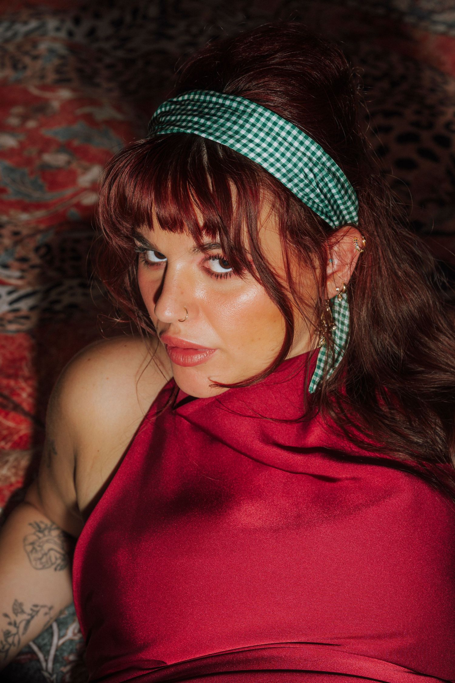 Woman in a red halter top with a green gingham headband and matching earrings, posing on a patterned surface.