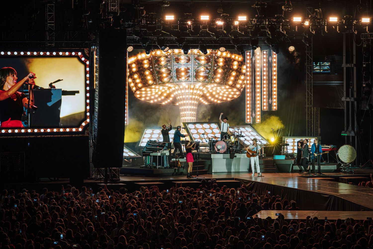 Large concert stage with full band performing under a spectacular crown-shaped chandelier light fixture.