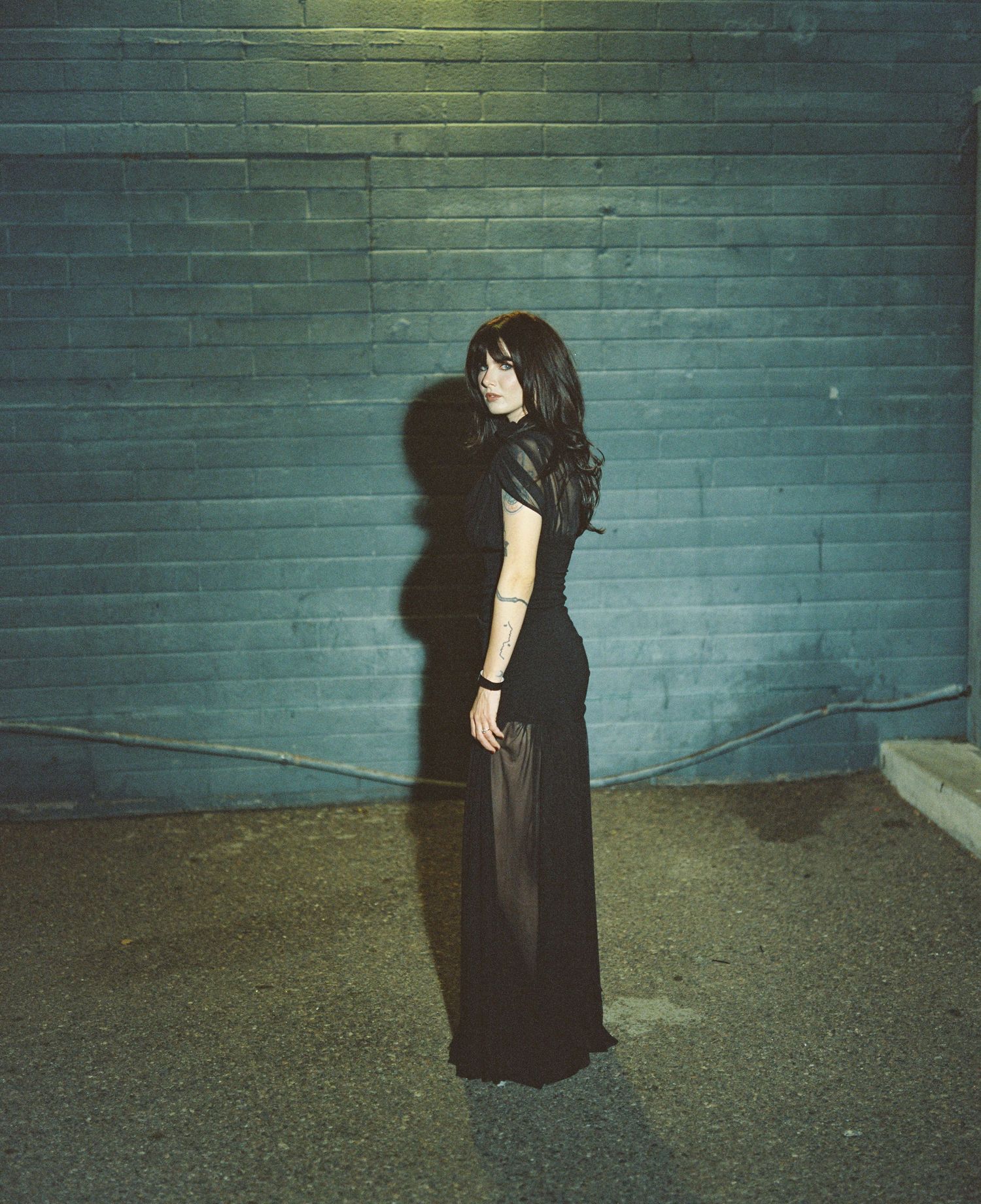 Woman in black dress standing against teal brick wall at night casting shadow.