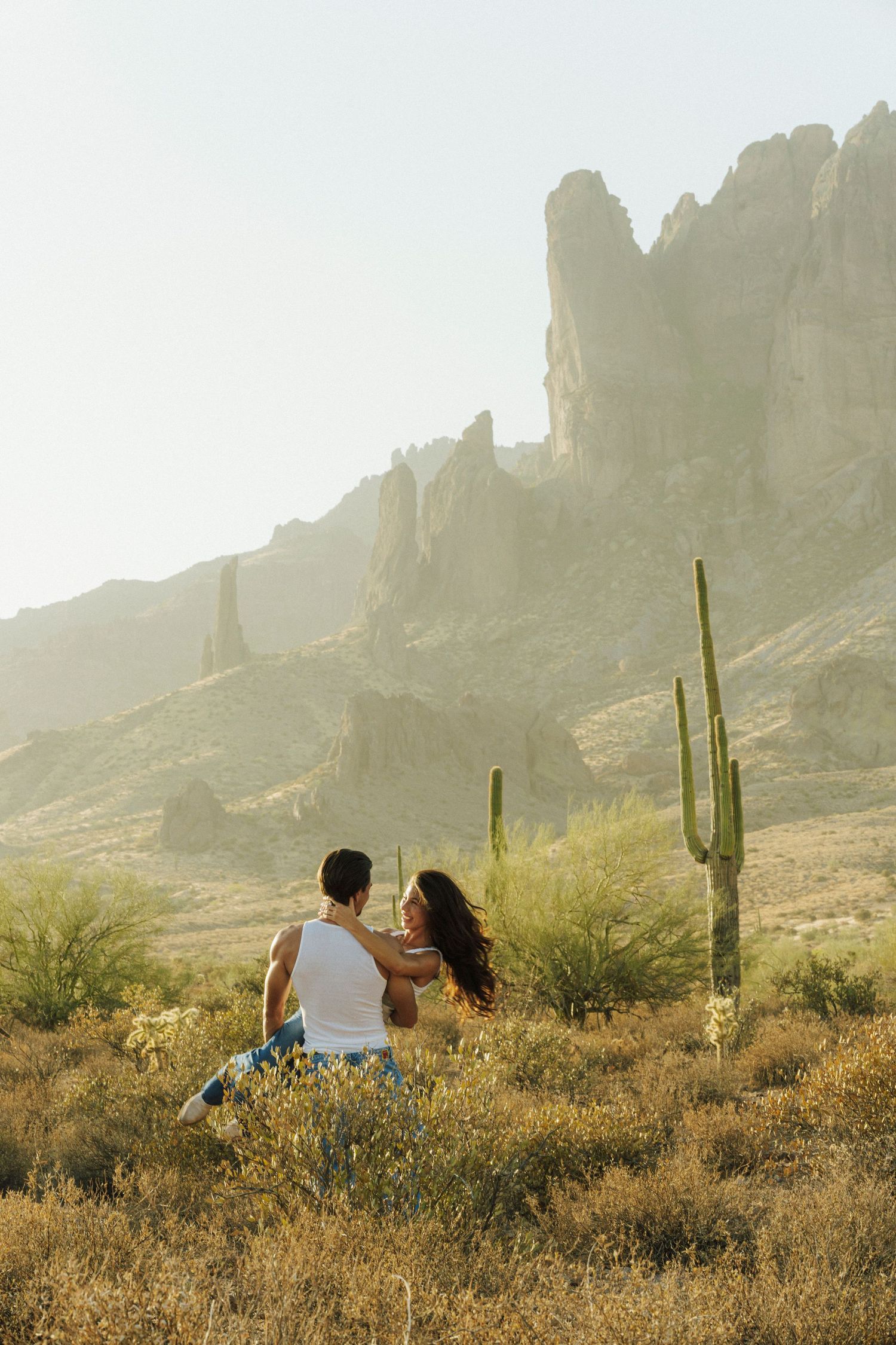 Couple embracing in Arizona desert landscape with saguaro cactus and Superstition Mountains in golden sunlight.