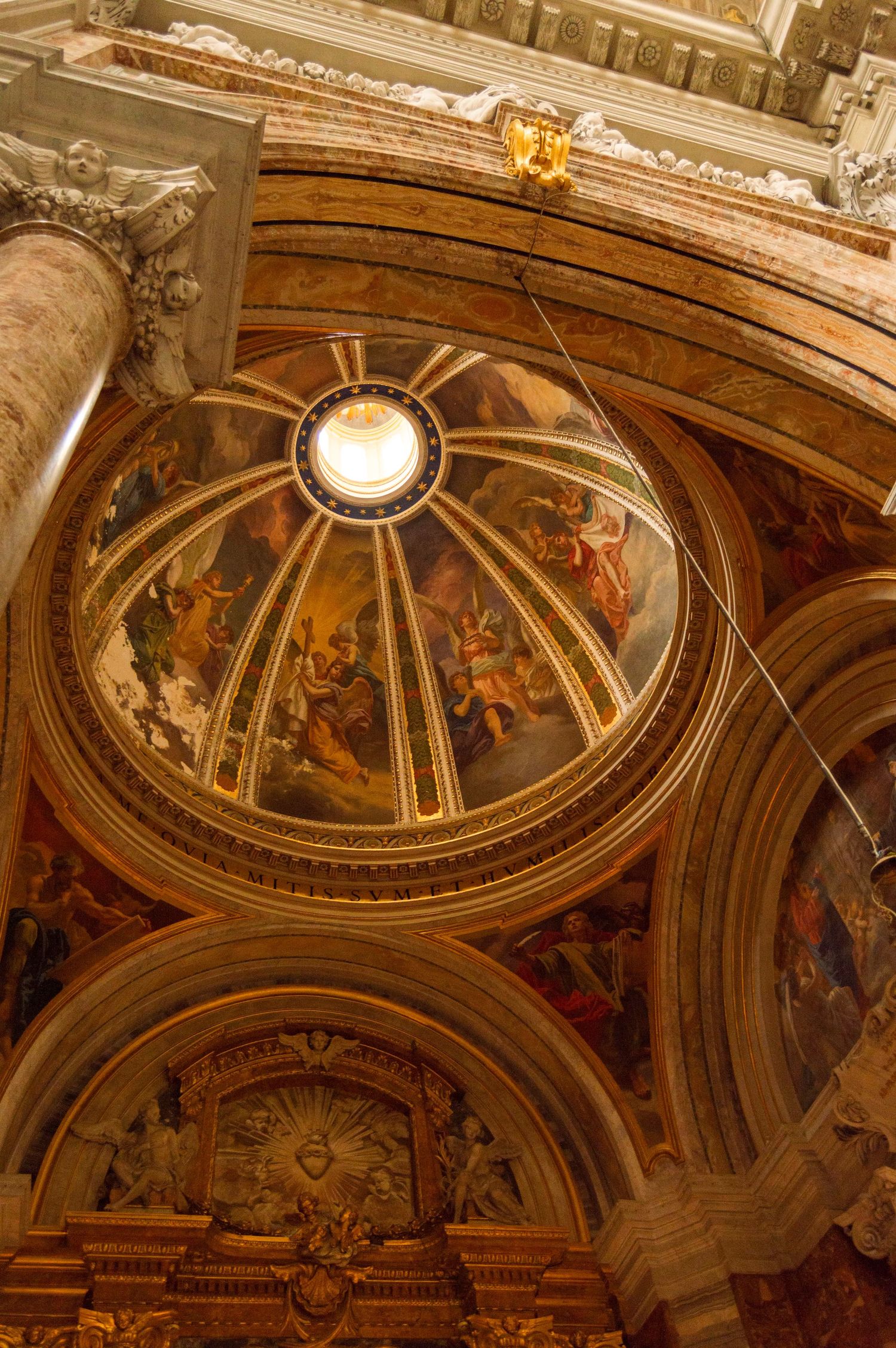 Ornate painted baroque cathedral dome interior with golden details and religious frescoes illuminated by skylight.