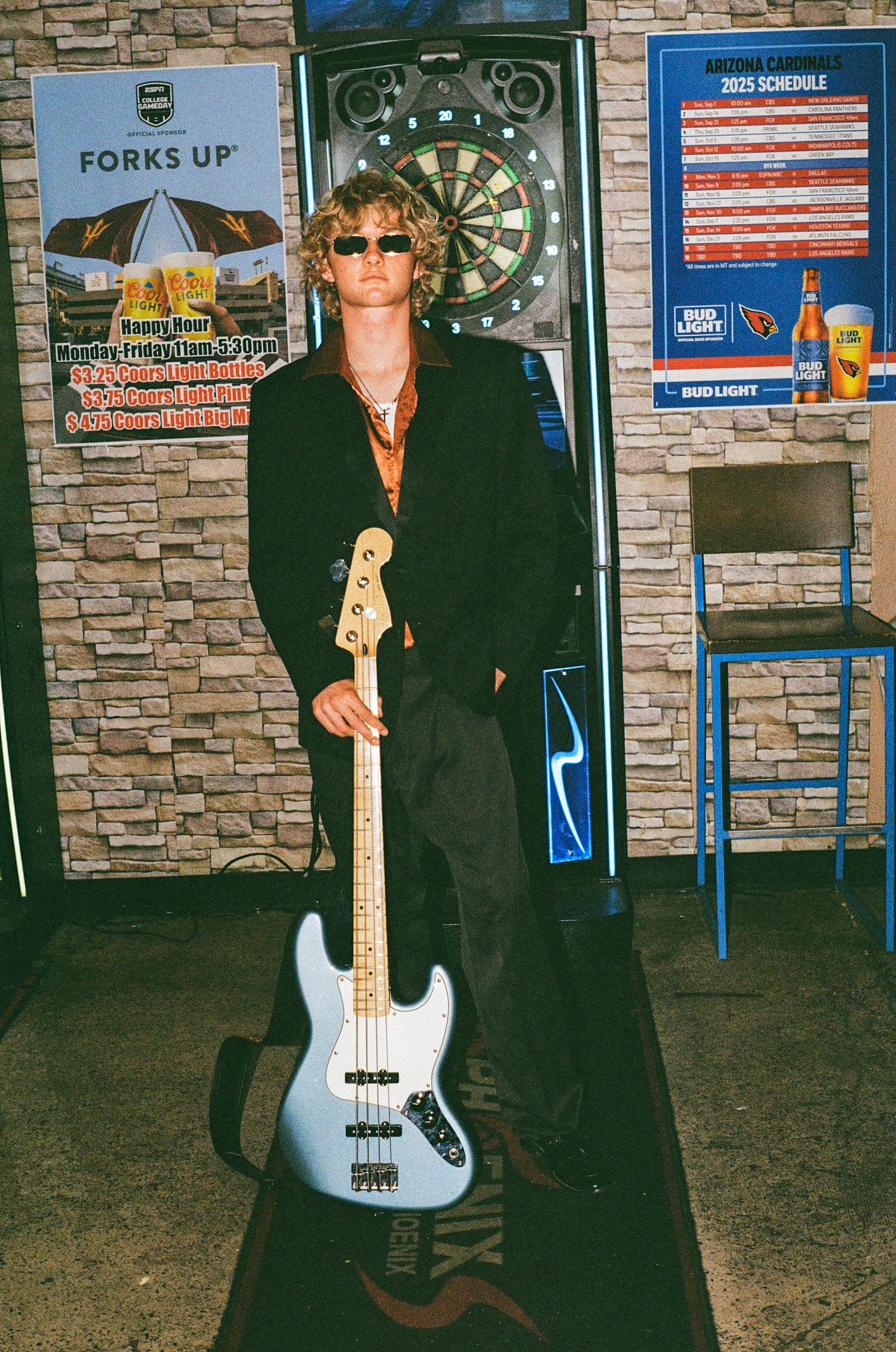 Young musician with curly hair and sunglasses holding a light blue bass guitar in a bar with dartboard.