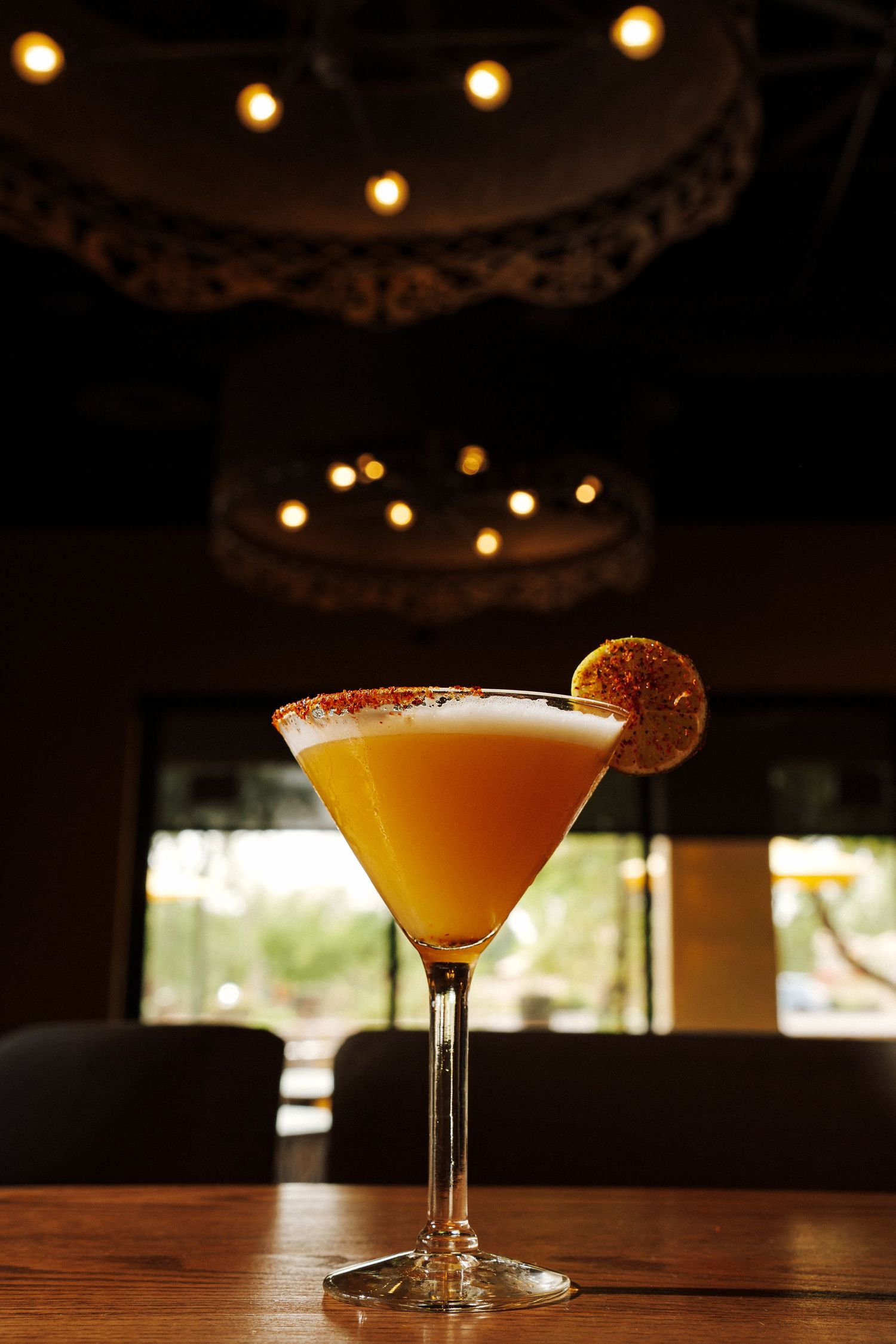 Golden orange martini cocktail with frothy top, citrus garnish, and salted rim in dimly lit bar setting.
