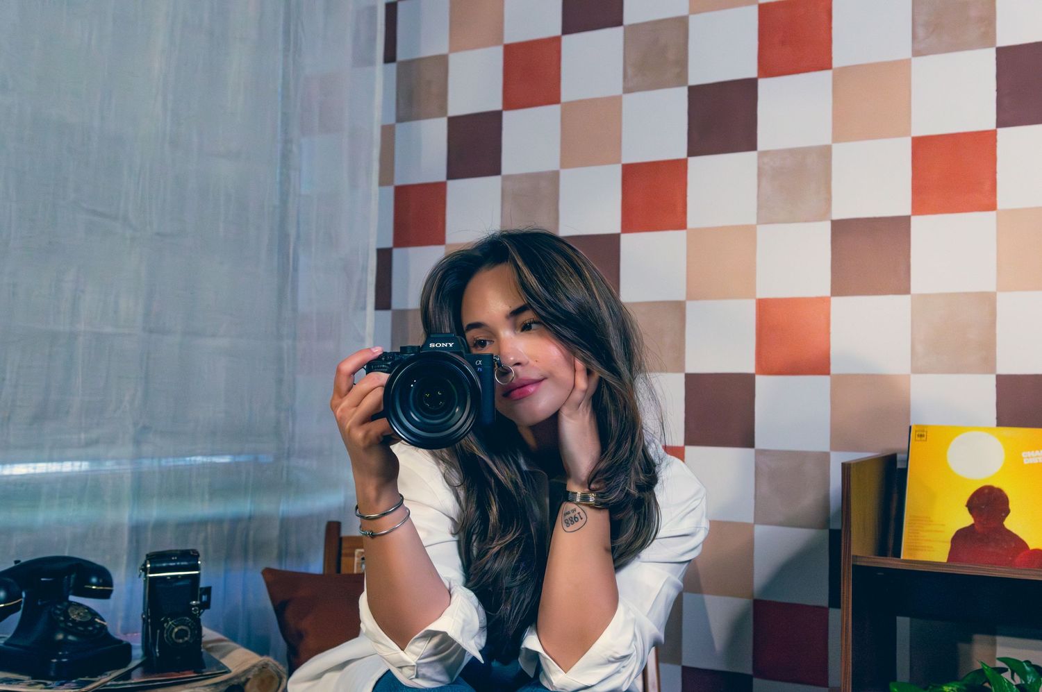 Young woman holding a camera in a retro-styled room with colorful checkered wall tiles.