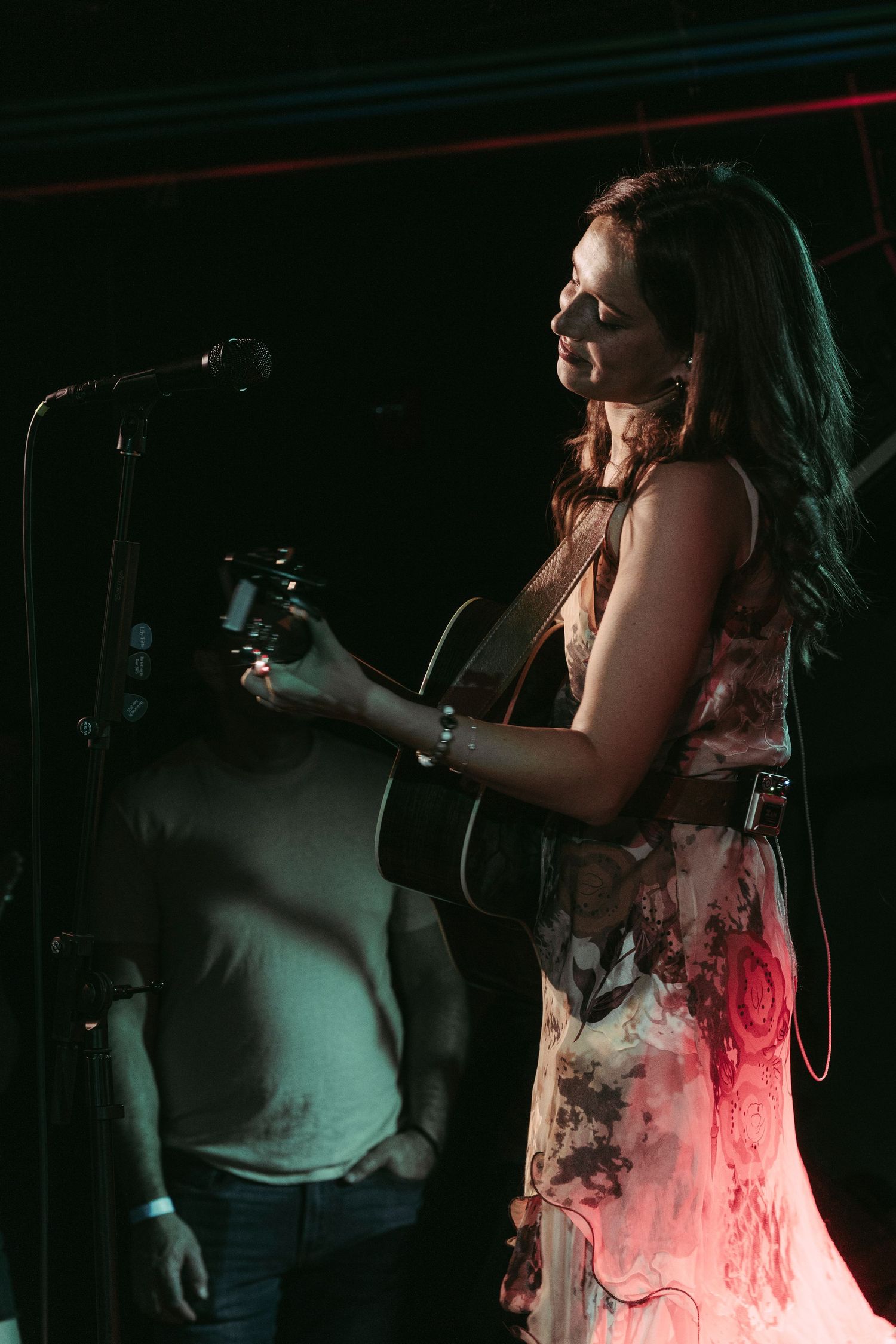Woman in a floral dress playing acoustic guitar and singing into a microphone on a dimly lit stage.