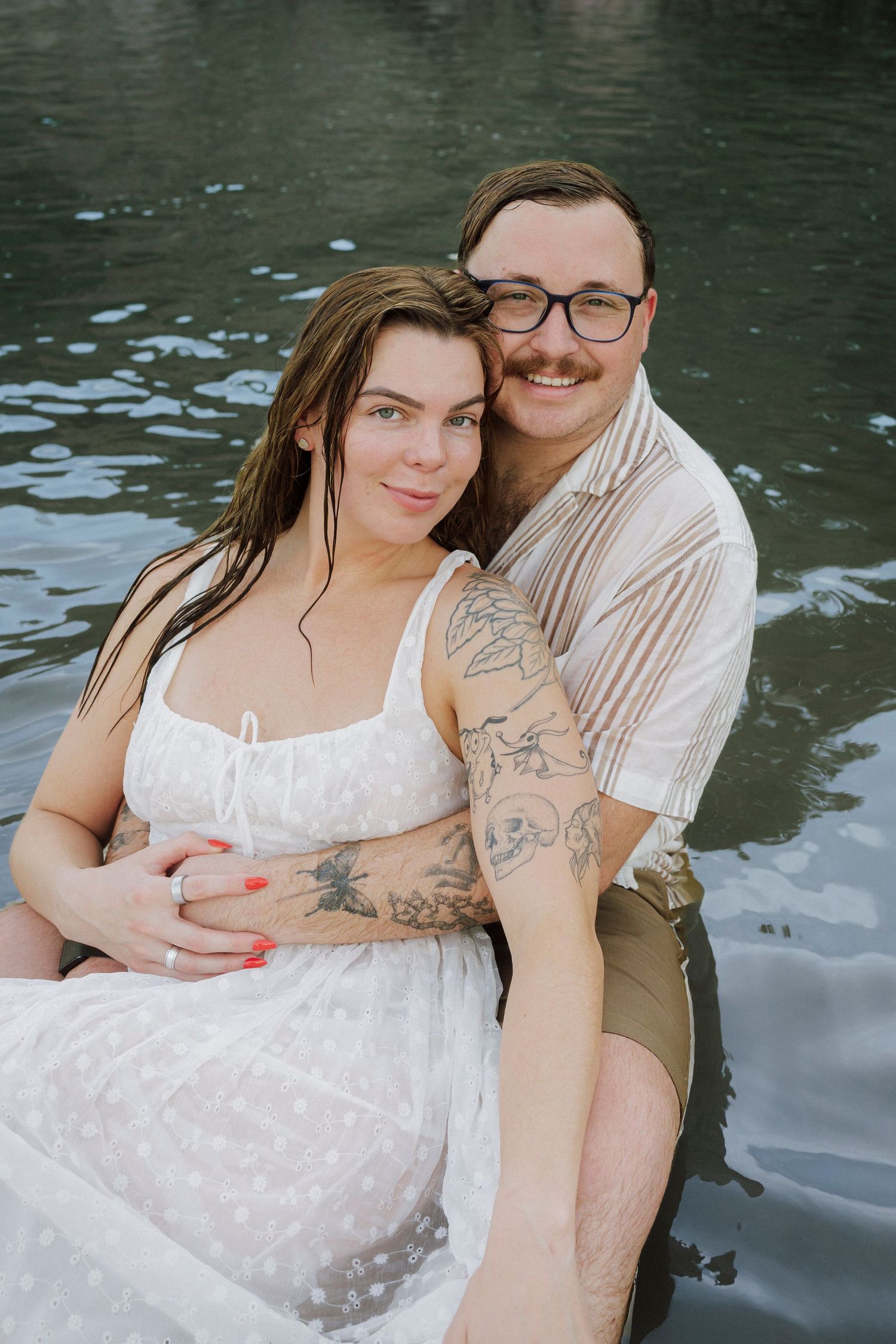 A couple sits together in water, woman in white dress with wet hair, man with glasses and tattoos embracing her tenderly.