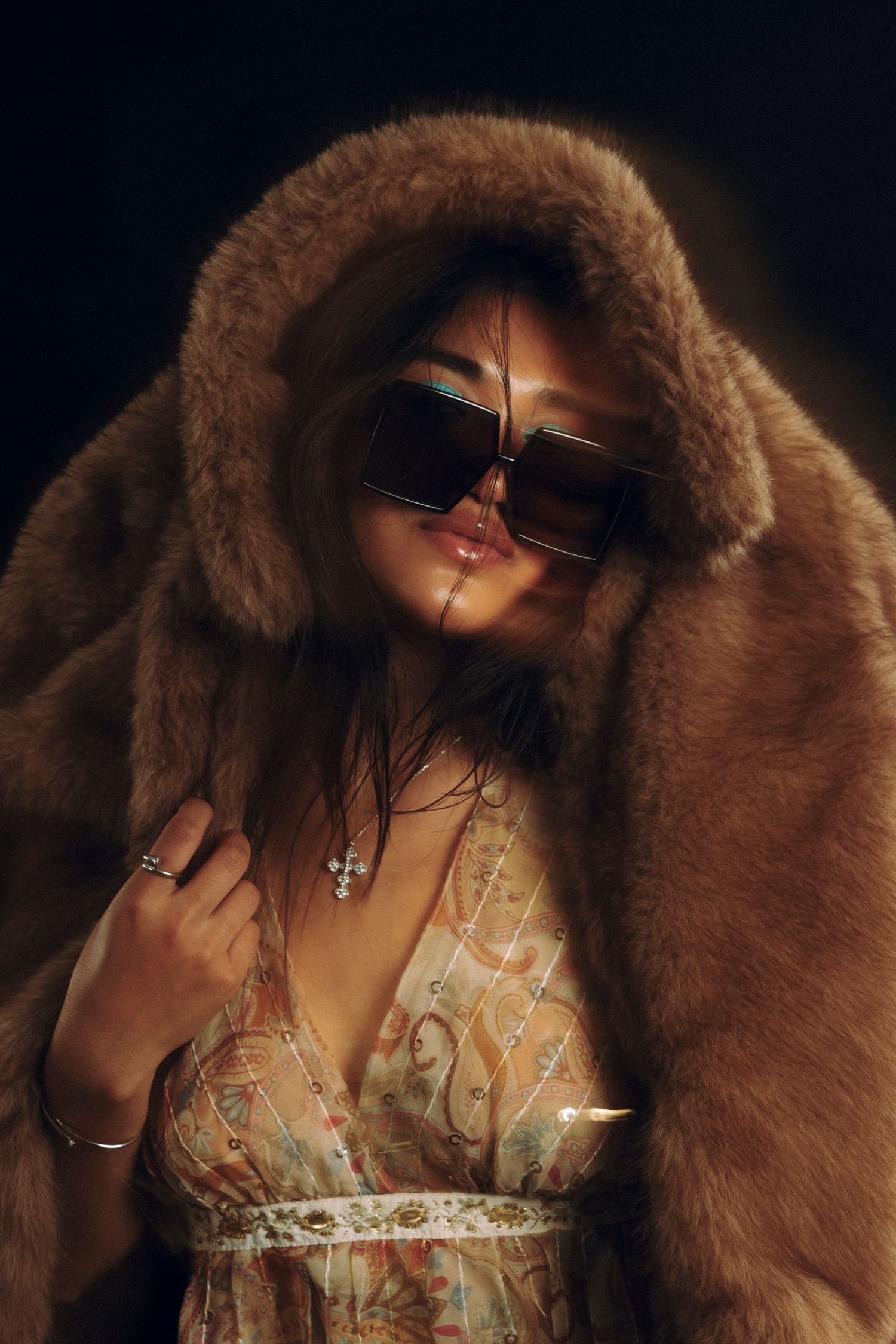 Woman in brown fur coat and oversized sunglasses wearing floral dress and jewelry in dramatic lighting.