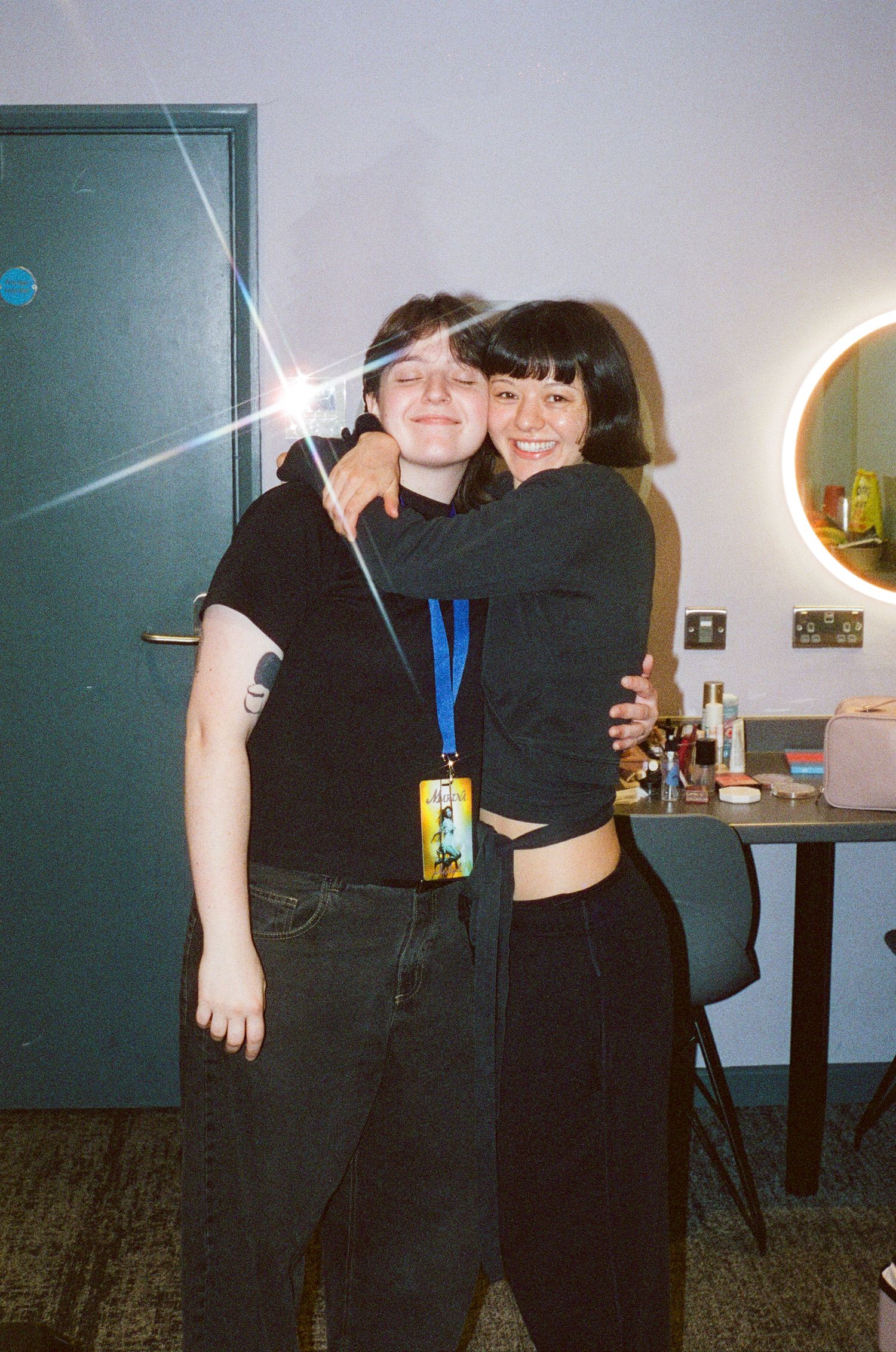 Two young women in black outfits hugging and smiling in a room with a round mirror and vanity table.