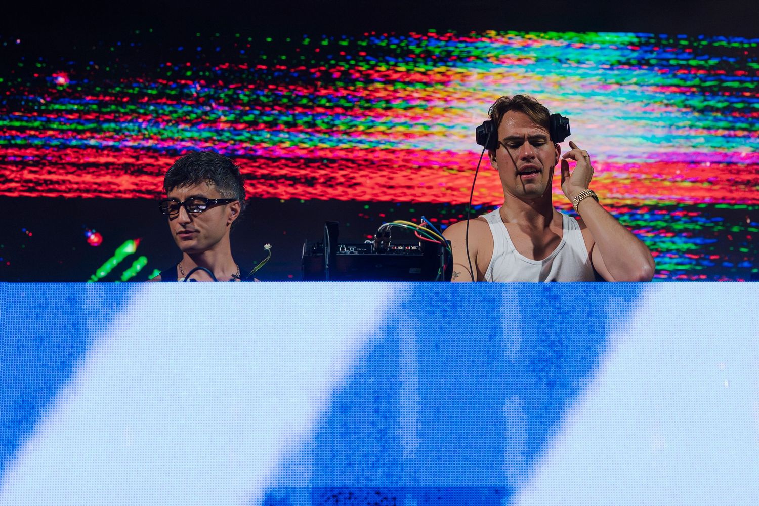 Two DJs performing at a vibrant music festival, one wearing headphones behind a DJ booth with colorful LED lights.