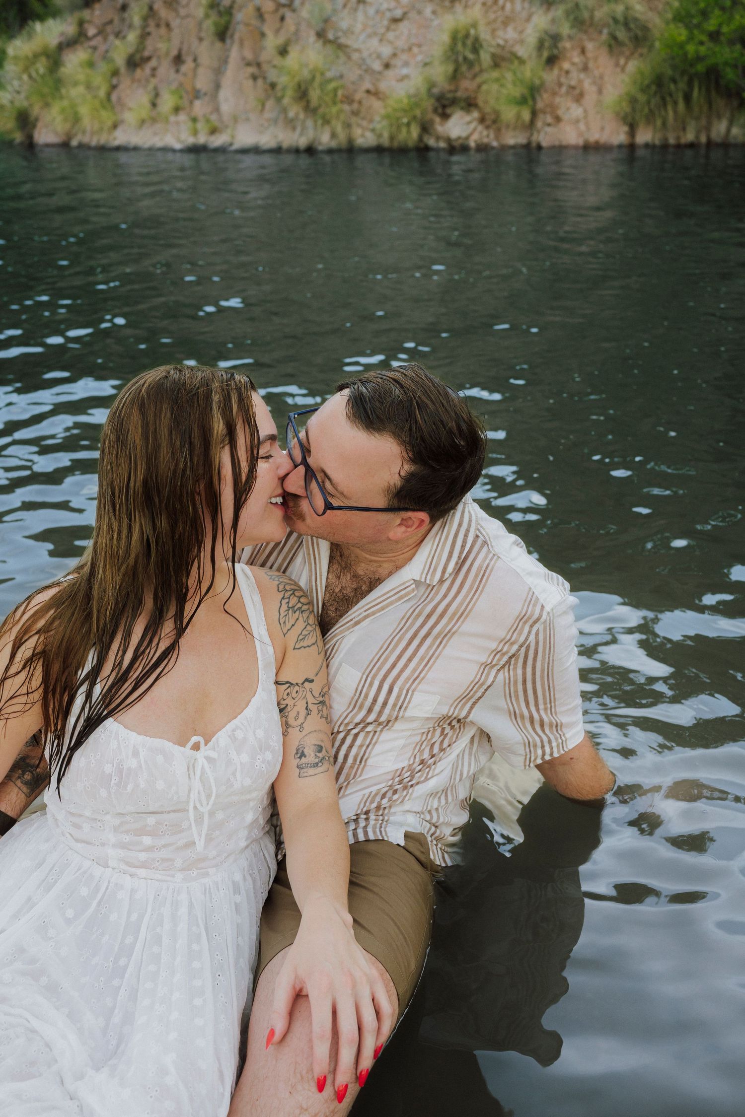 Couple sharing a romantic kiss while sitting in a serene lake, woman in white dress with tattoos, man in striped shirt.