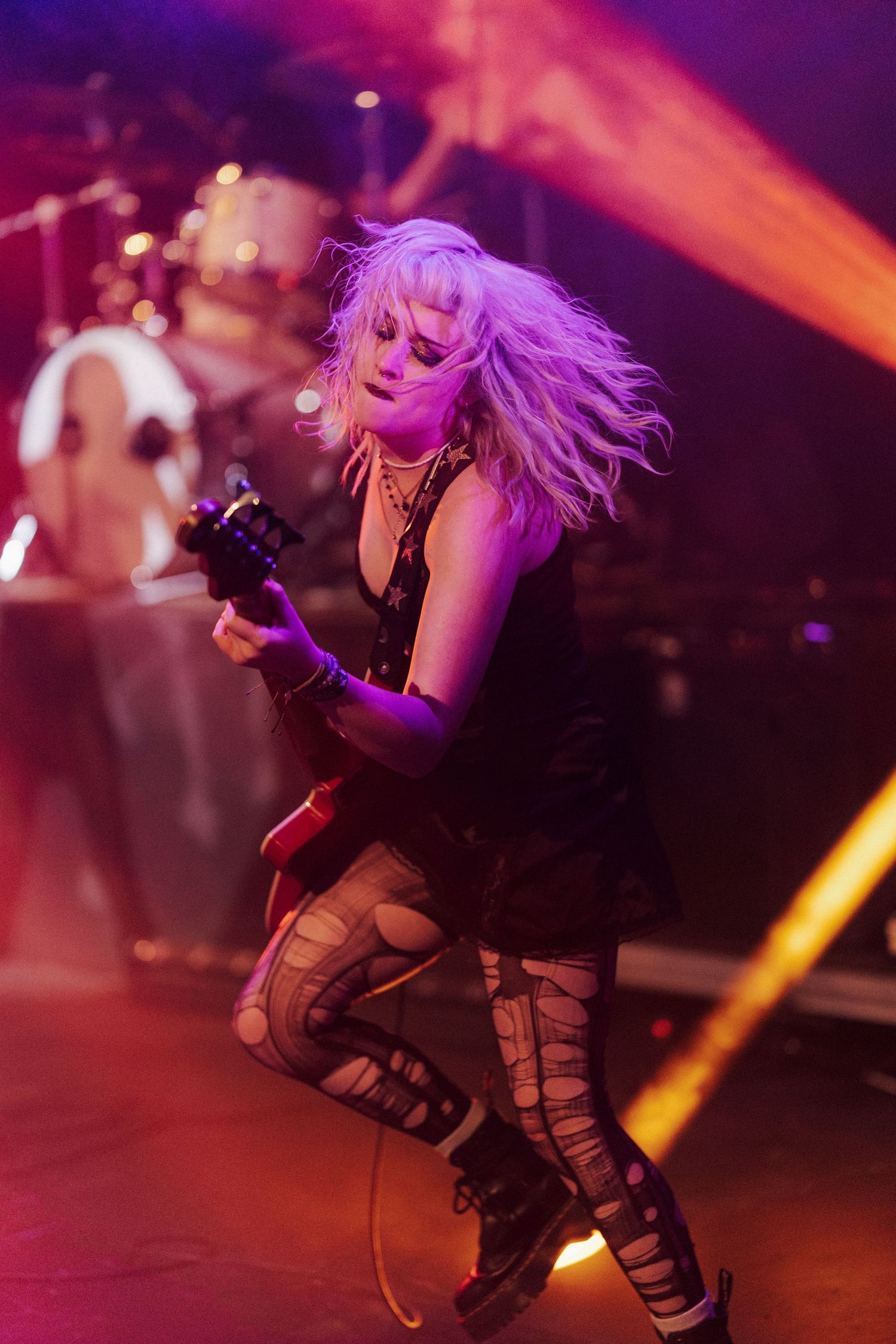 Blonde female guitarist performing energetically on stage under purple and orange concert lights.