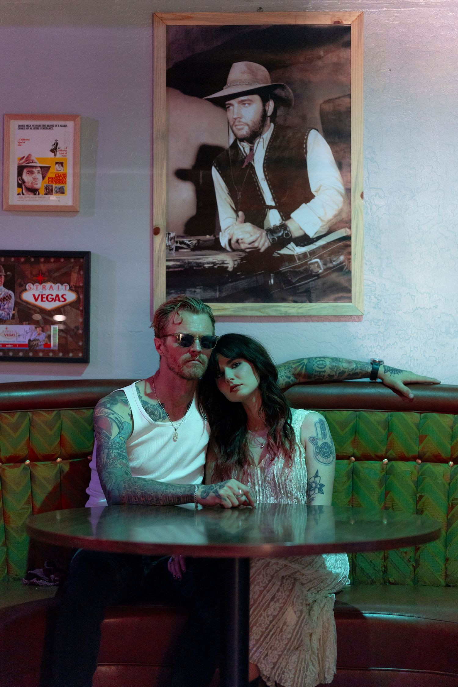 Tattooed couple sitting at a round table in a retro diner booth beneath a cowboy portrait and Vegas sign. From 'Dead Ends' music video