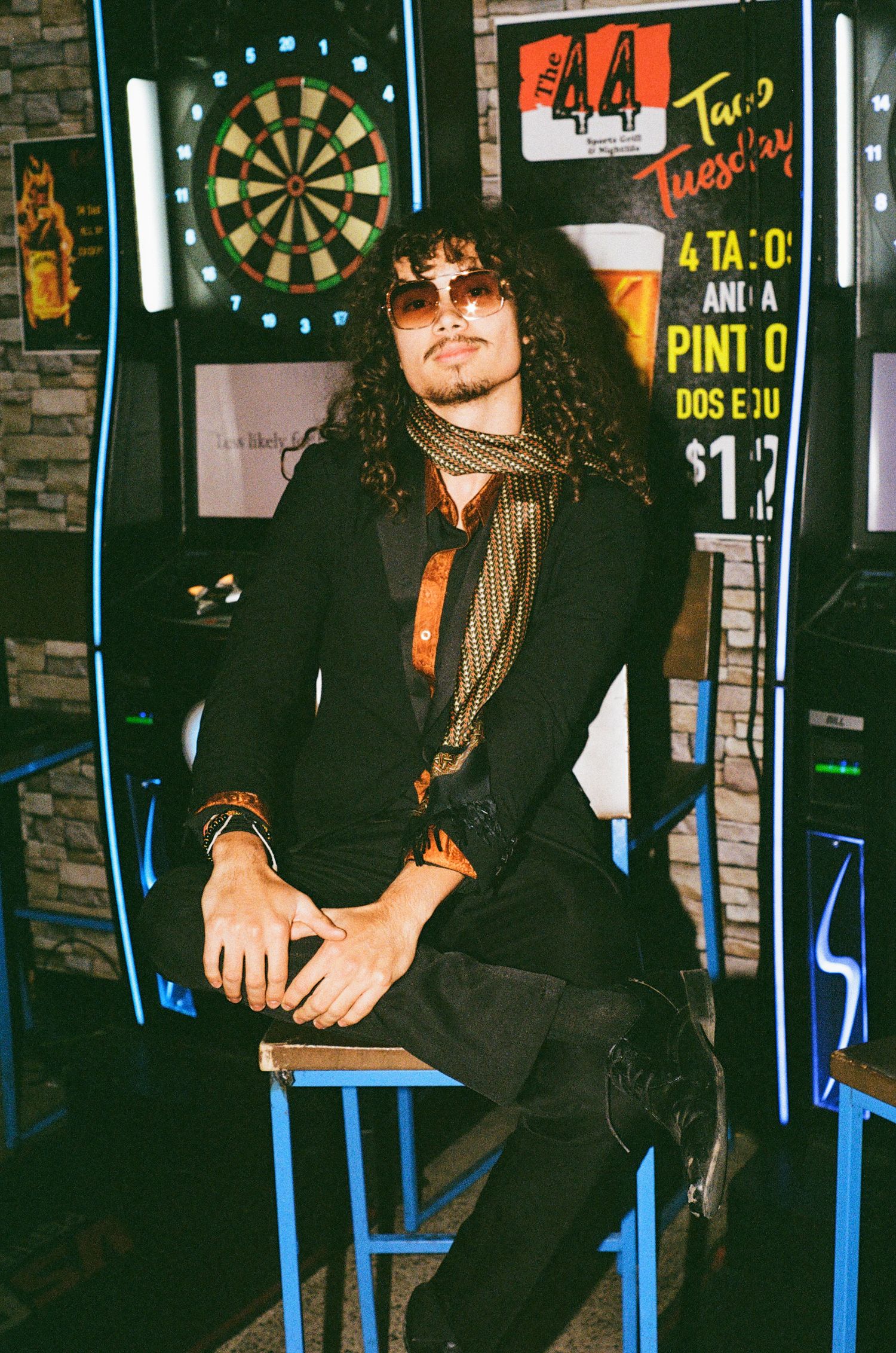 A stylish man with curly hair and sunglasses sits in a bar near a dartboard, wearing a black jacket and scarf.