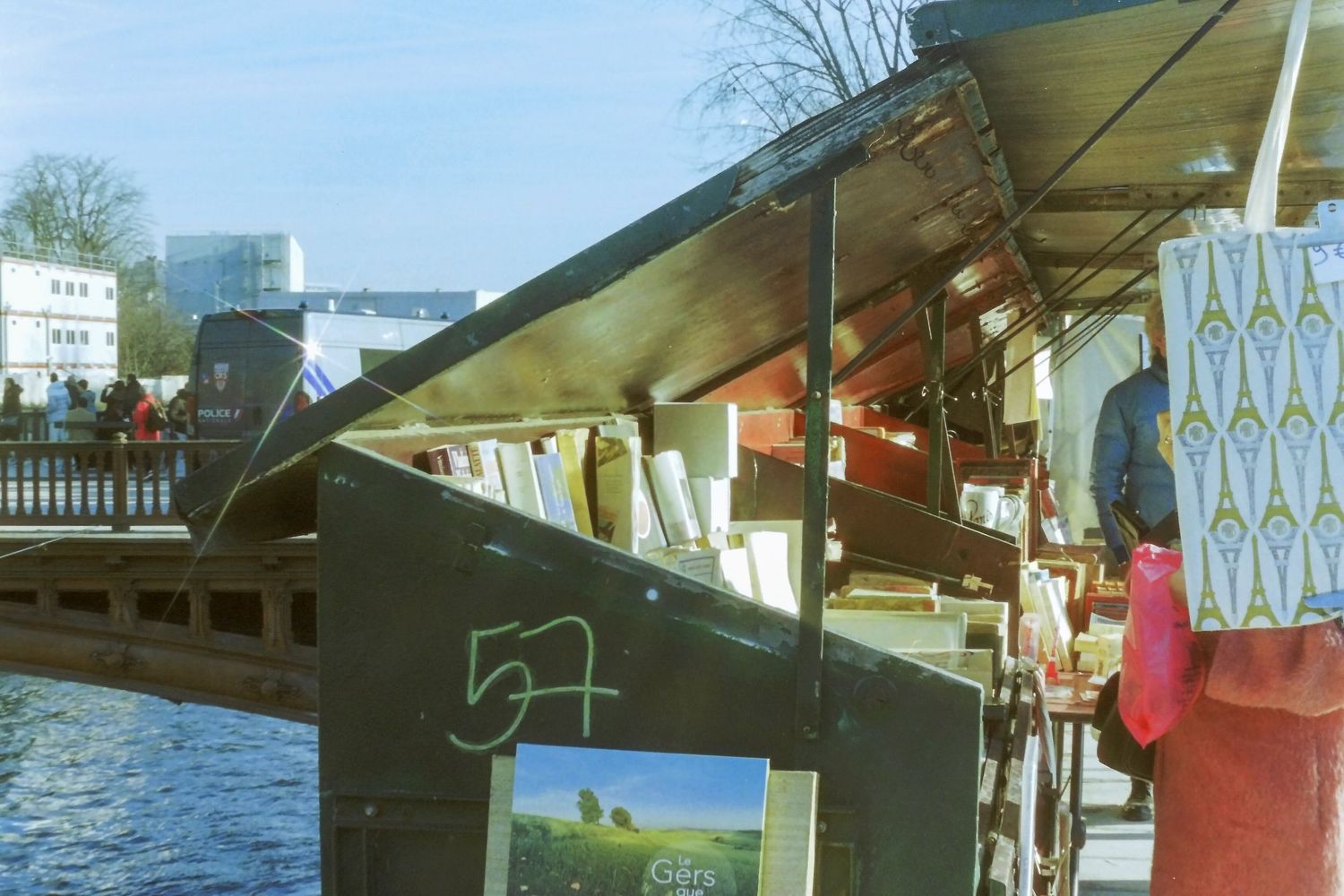 Outdoor book stall along a riverside canal with colorful prints and artwork displayed under a metal awning.