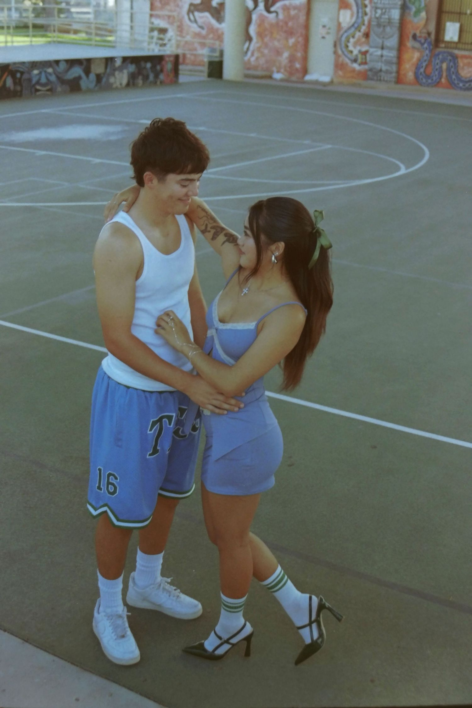 Young couple sharing a tender moment on a basketball court, man in blue shorts, woman in blue dress.