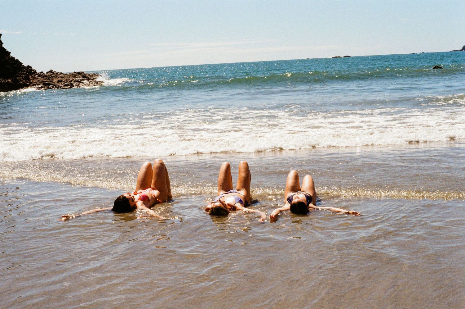 Three people lying in shallow ocean waves on a sunny beach with rocky coastline in background.