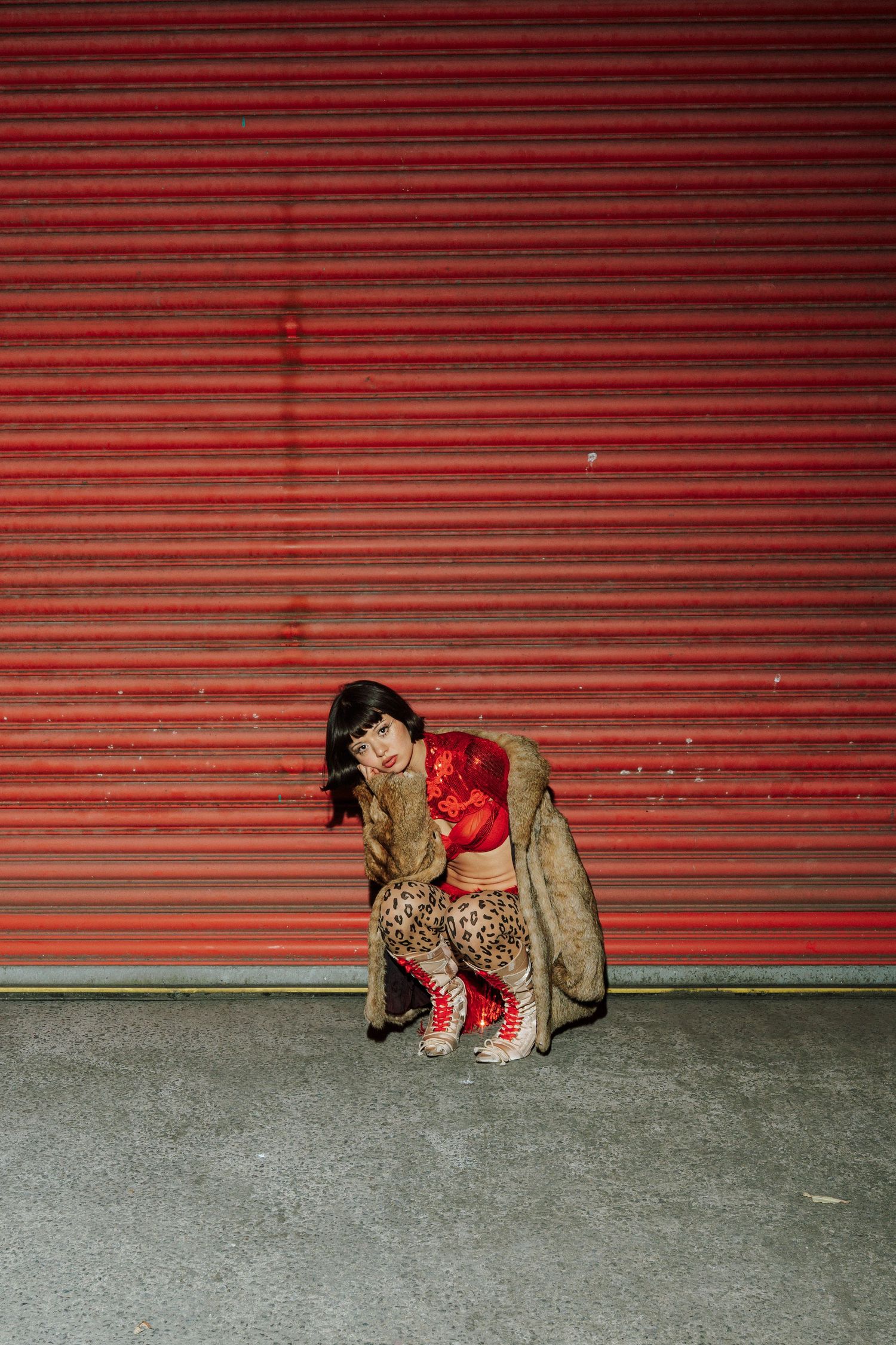 Woman in leopard pants, red top, fur coat, and red sneakers crouching in front of a red corrugated metal wall.