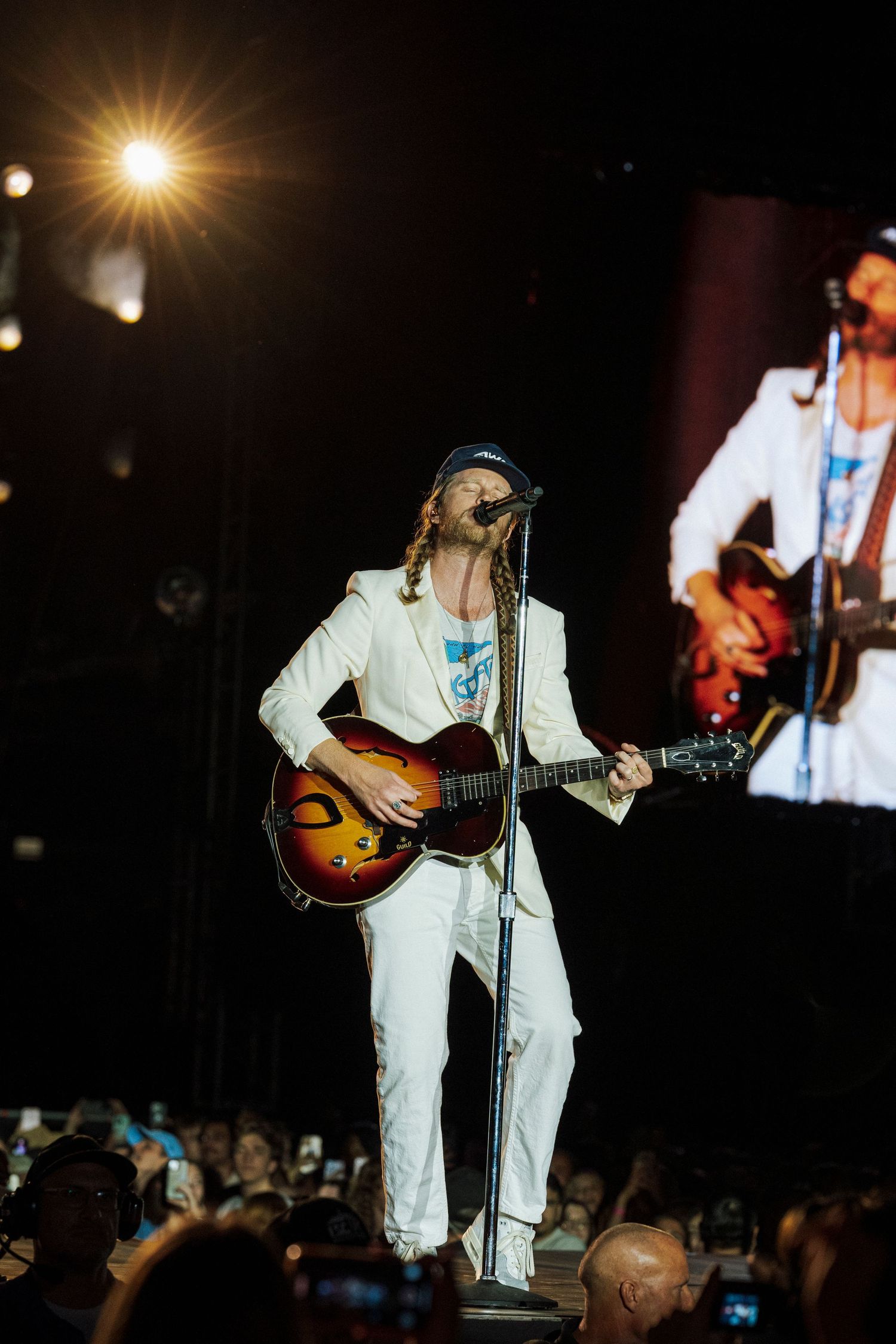 Guitarist performing live in a white suit playing a sunburst acoustic guitar on a concert stage.