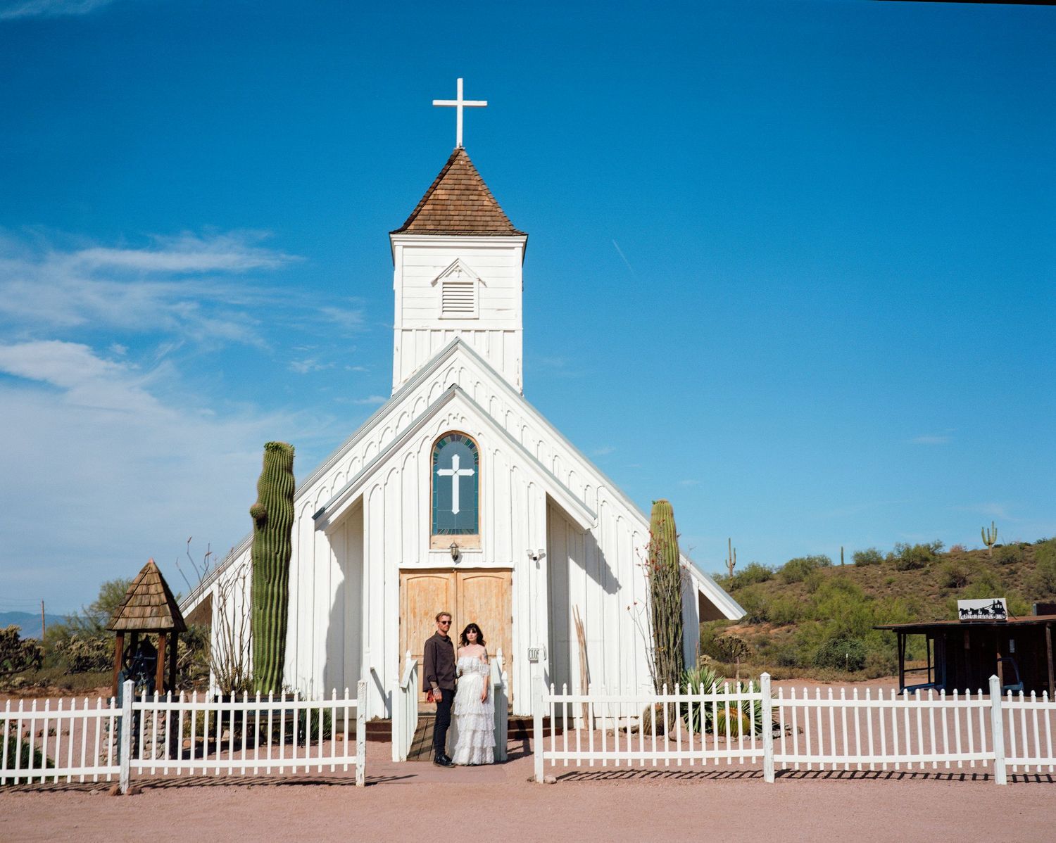 Couple posing in front of a white chapel with a cross at a desert wedding venue under a bright blue sky. From 'Dead Ends' music video