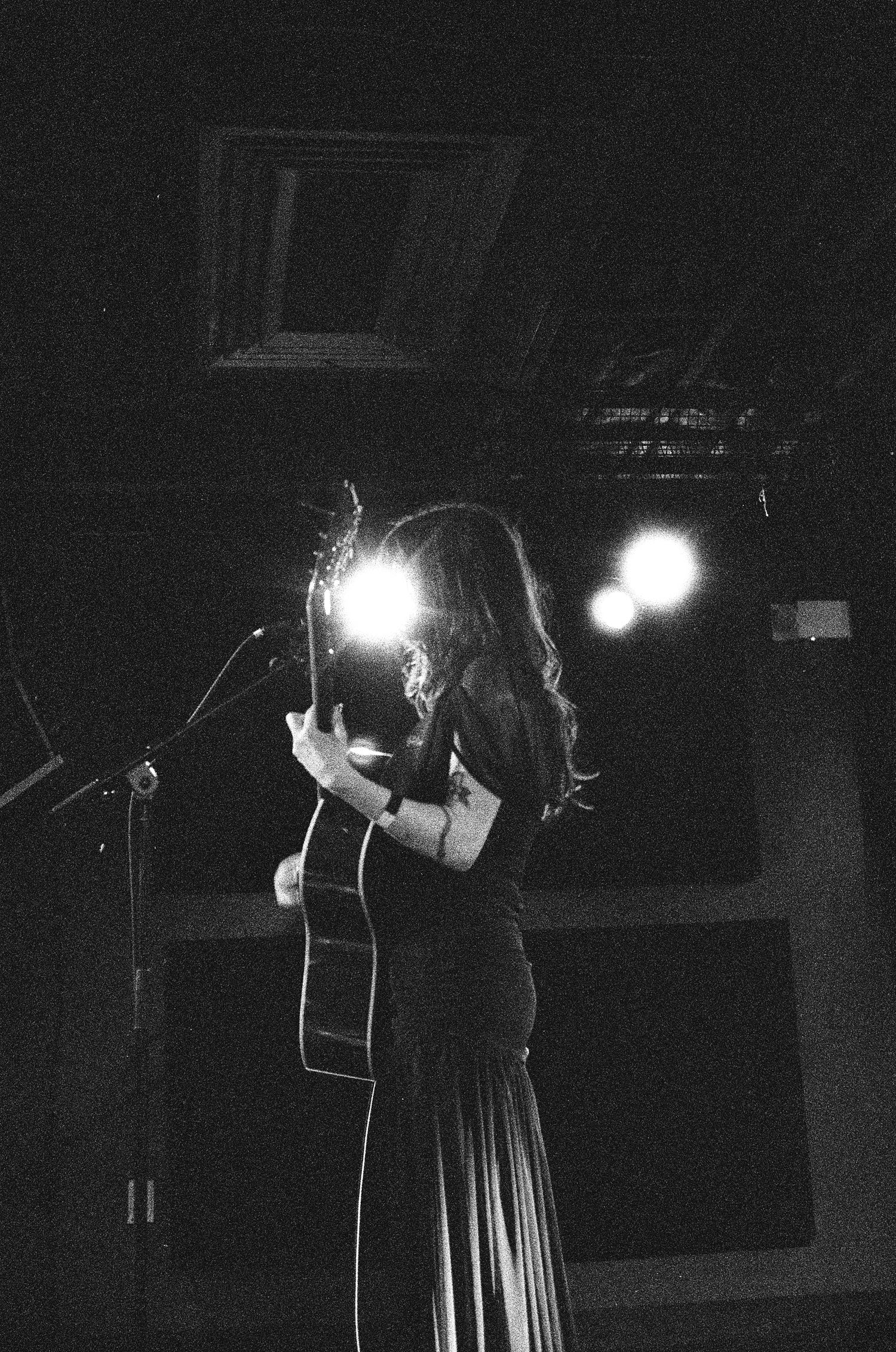 Black and white film photo of a woman holding an acoustic guitar on stage with bright backlighting.