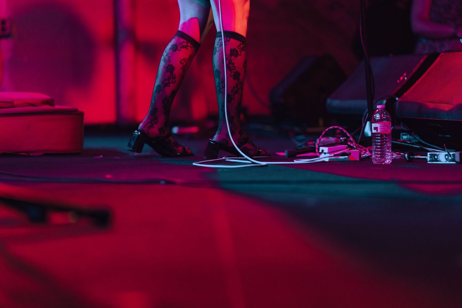Stage floor shot showing guitar pedals, cables, and a performer's legs under pink and red lighting.