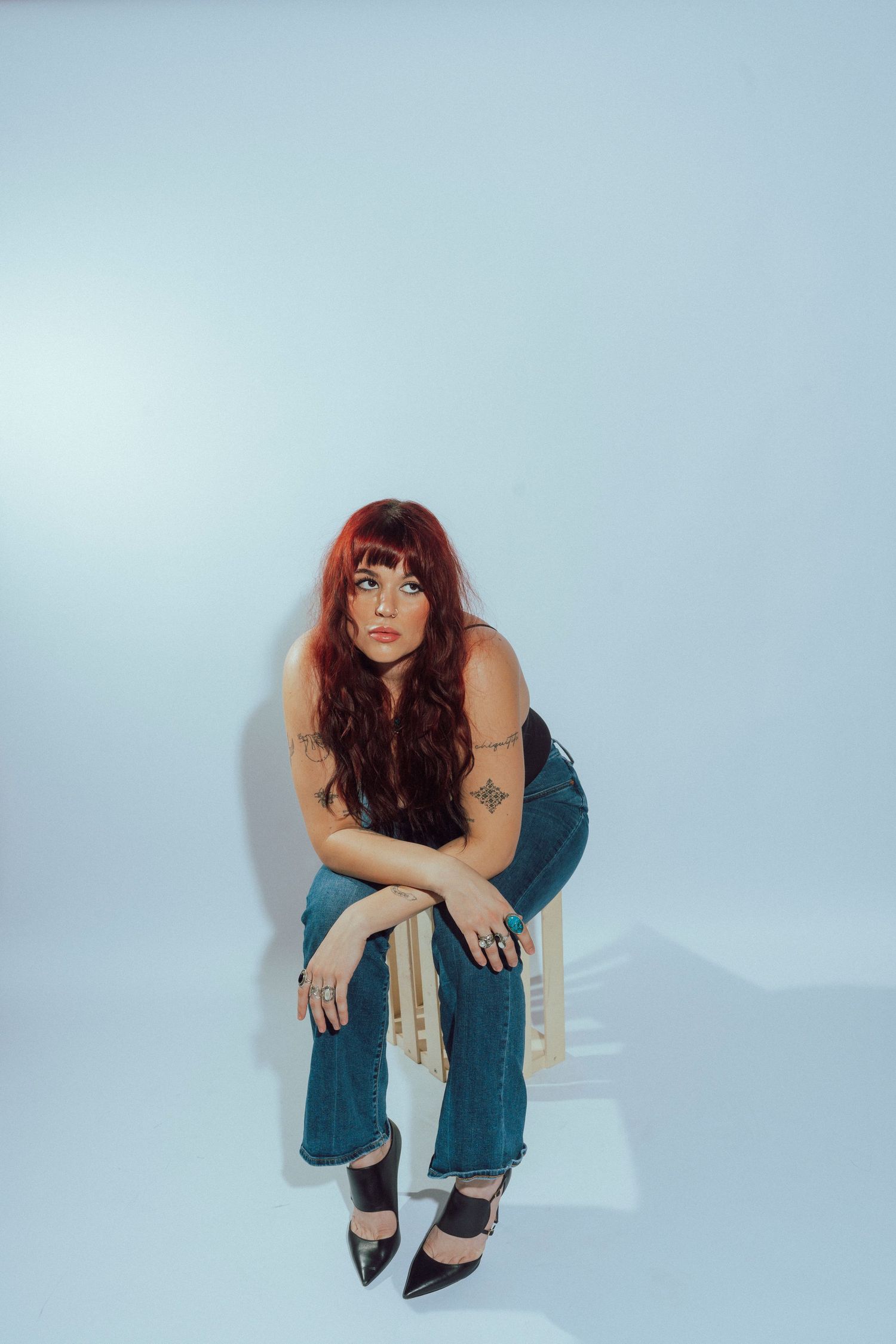 Red-haired woman with tattoos sits on wooden stool in jeans and black heels against light blue background.
