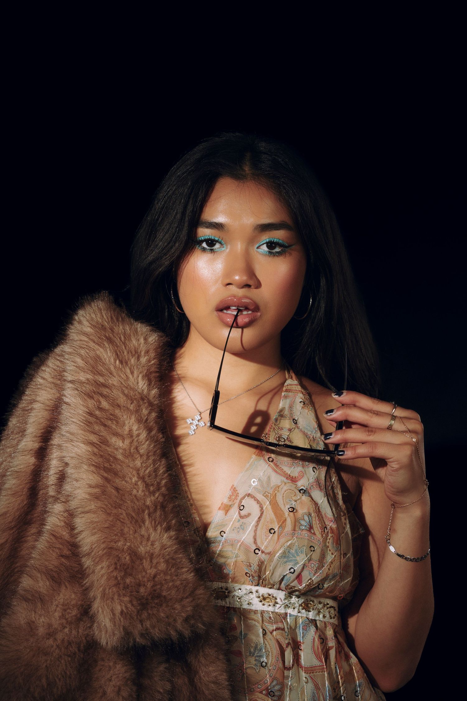 Young Asian woman in floral dress and brown fur stole holds sunglasses, posing against dark background.