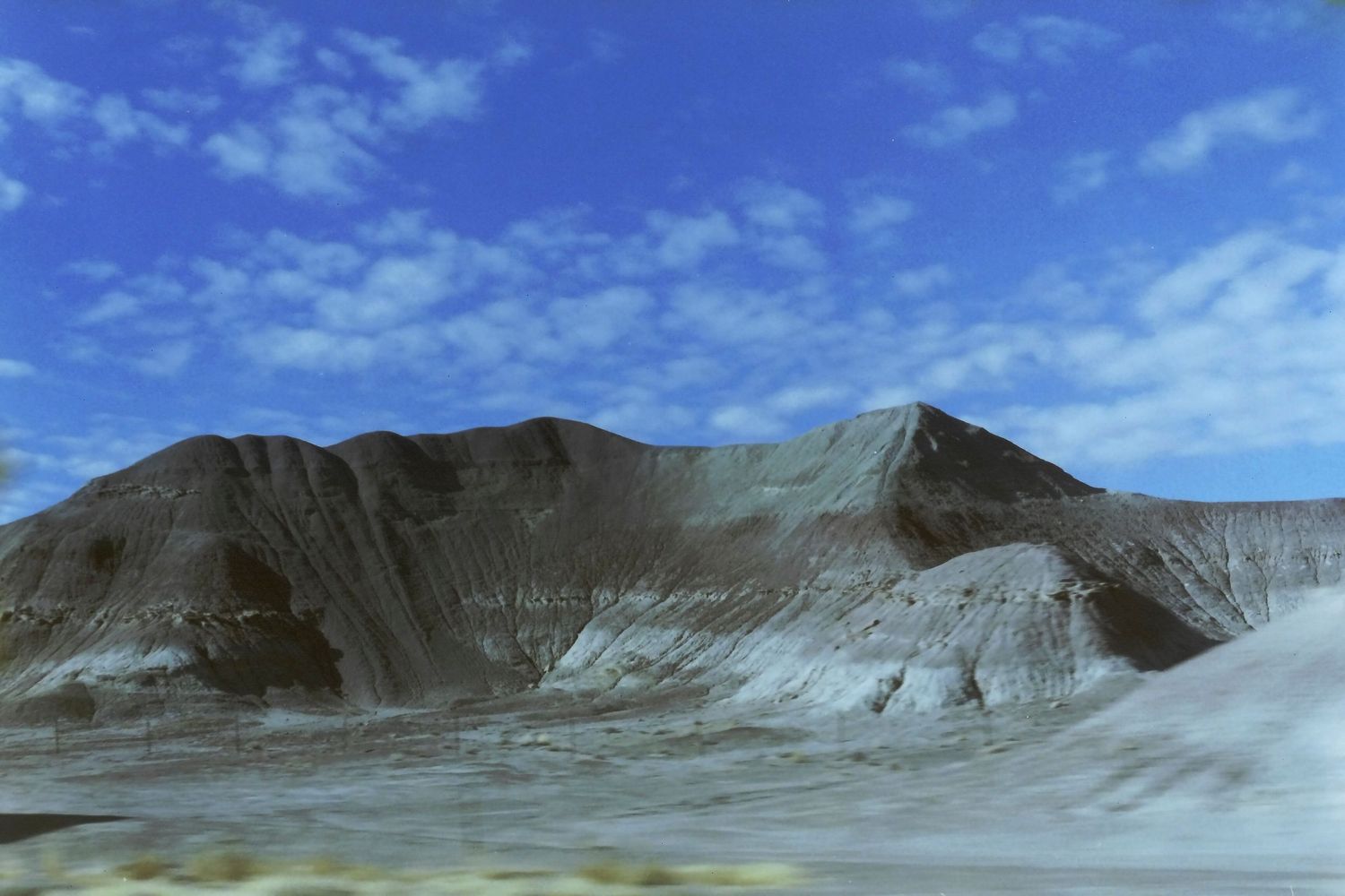 Arid desert landscape with a large eroded mountain formation under a vivid blue sky with wispy clouds.