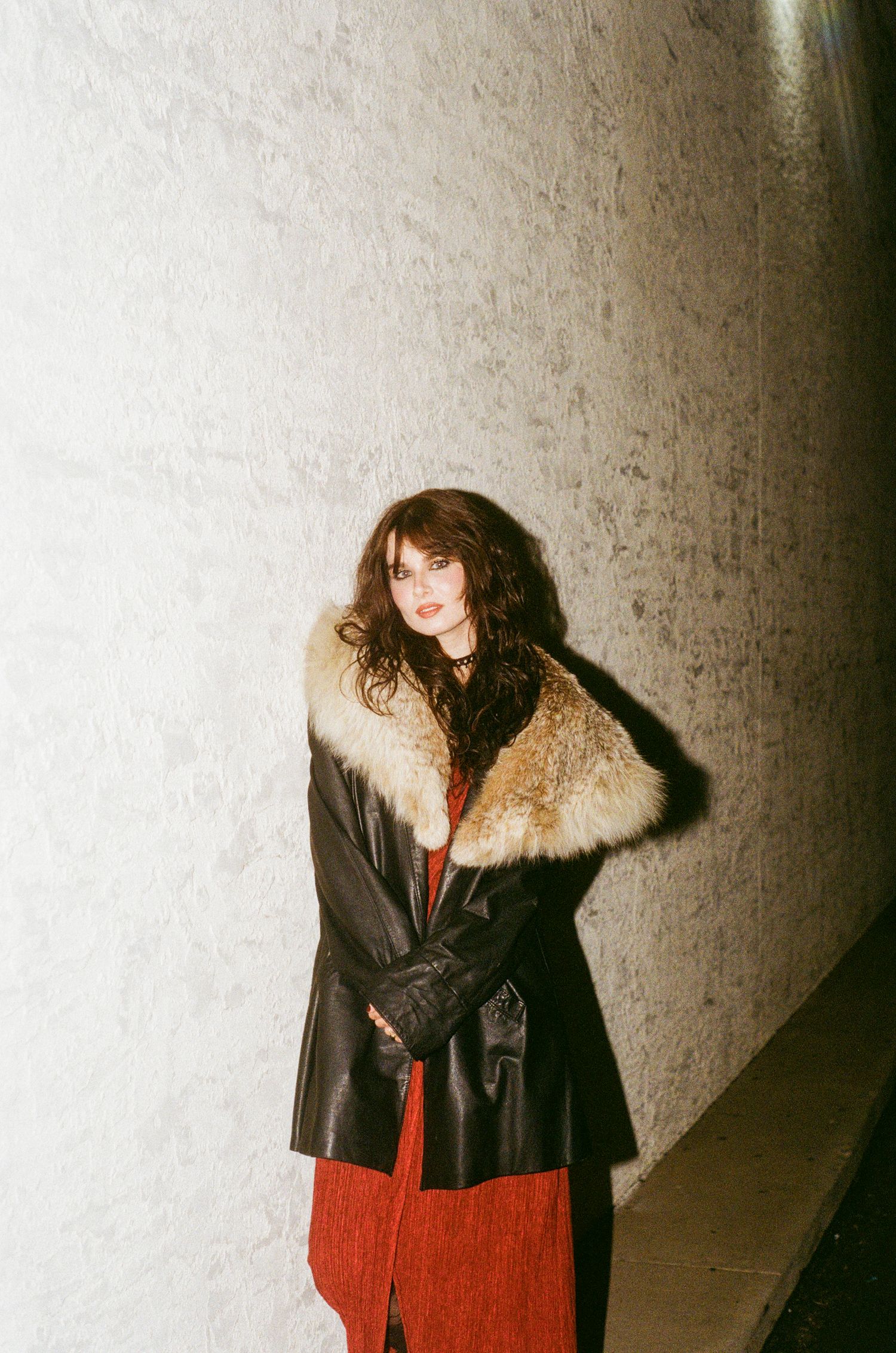 Woman in black leather coat with fur collar stands against concrete wall in film photography style.