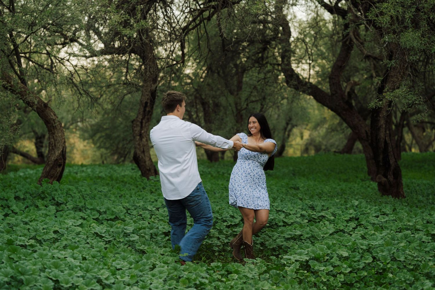 Joyful couple dancing and spinning in a lush green forest clearing surrounded by trees.