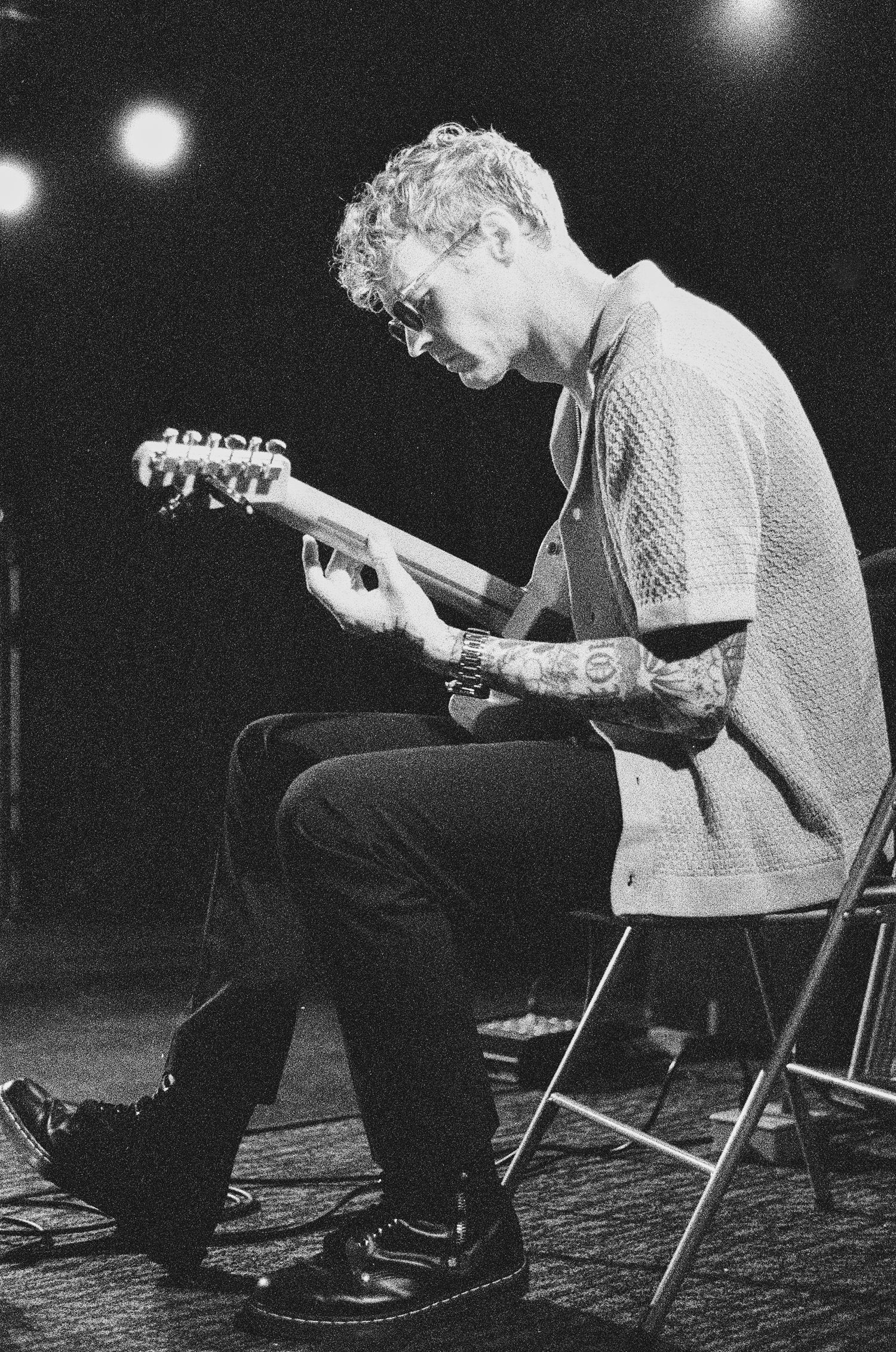 Black and white photo of a tattooed man sitting and playing electric guitar under stage lights.