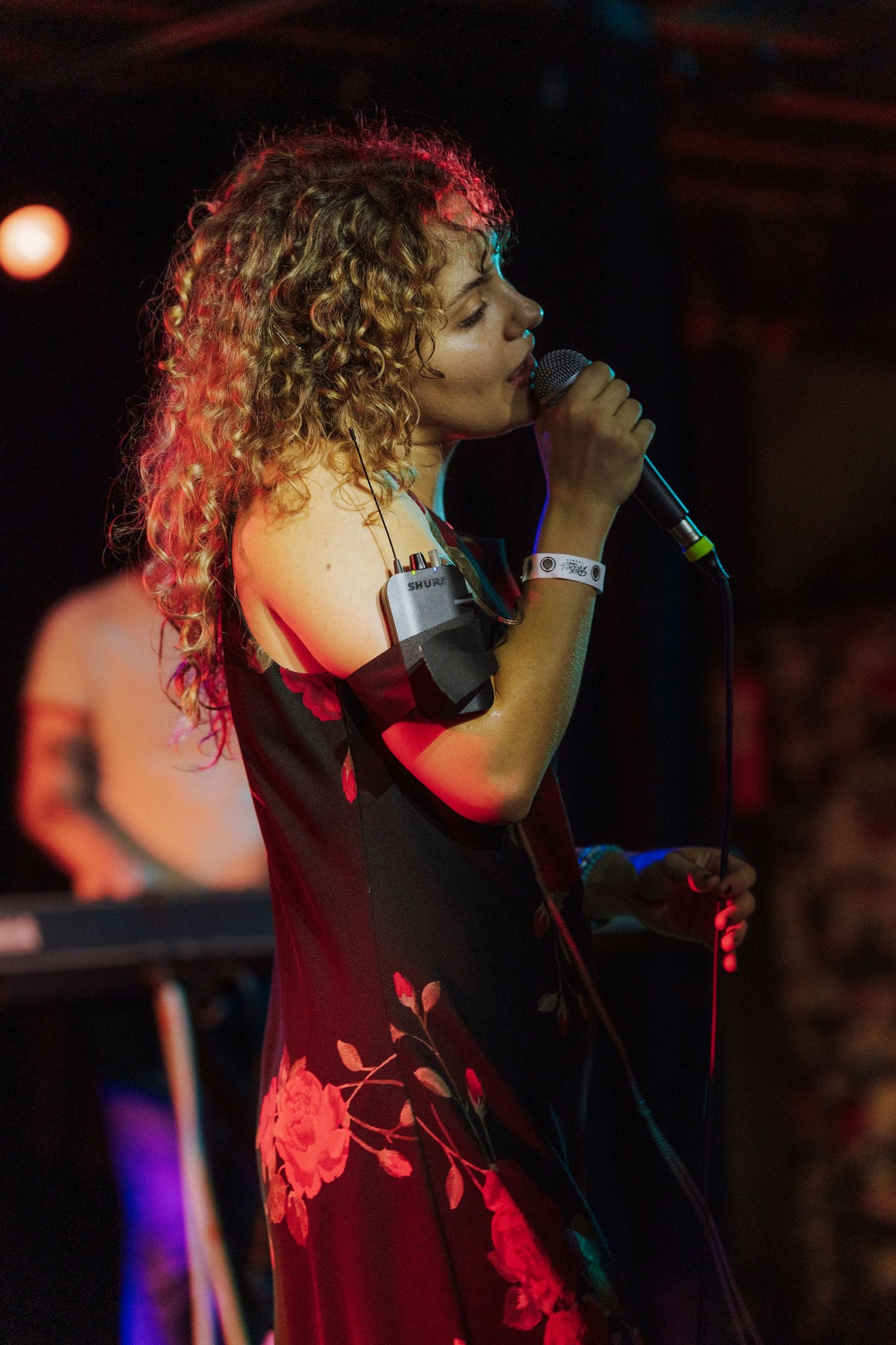 Curly-haired female vocalist in floral dress performs on stage under warm red concert lights.