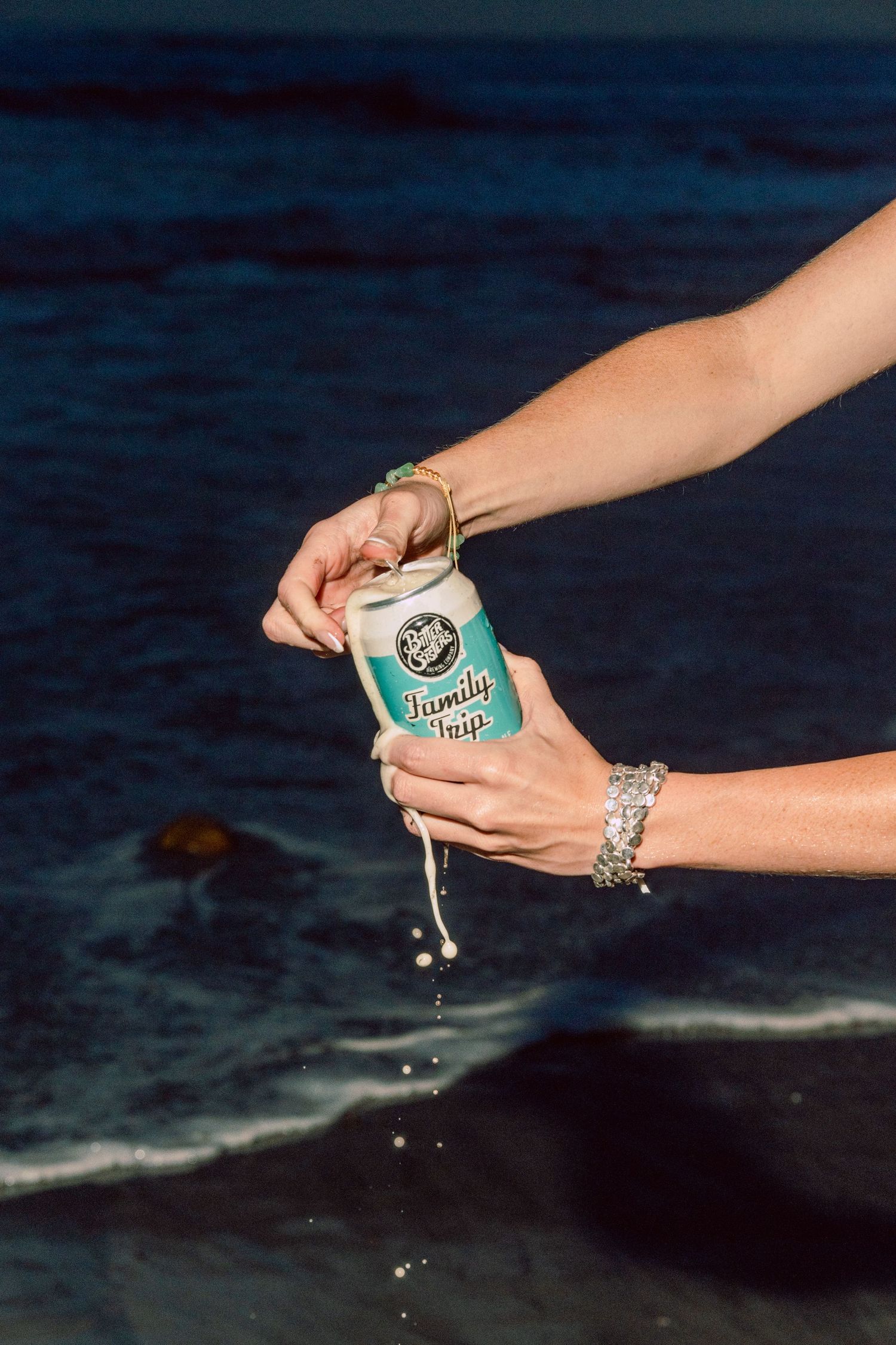 Hands with bracelets opening a Family Hops beer can over ocean waves at night, with liquid dripping down.