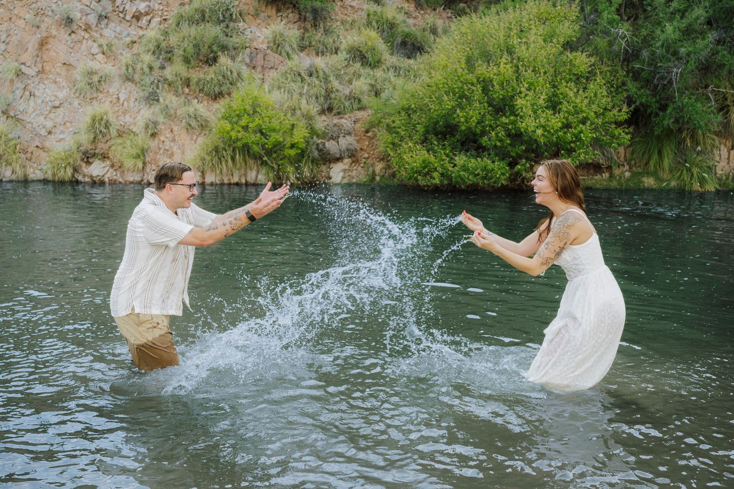 Couple splashing water at each other while standing in a river, wearing white outfits during a fun engagement photo session.