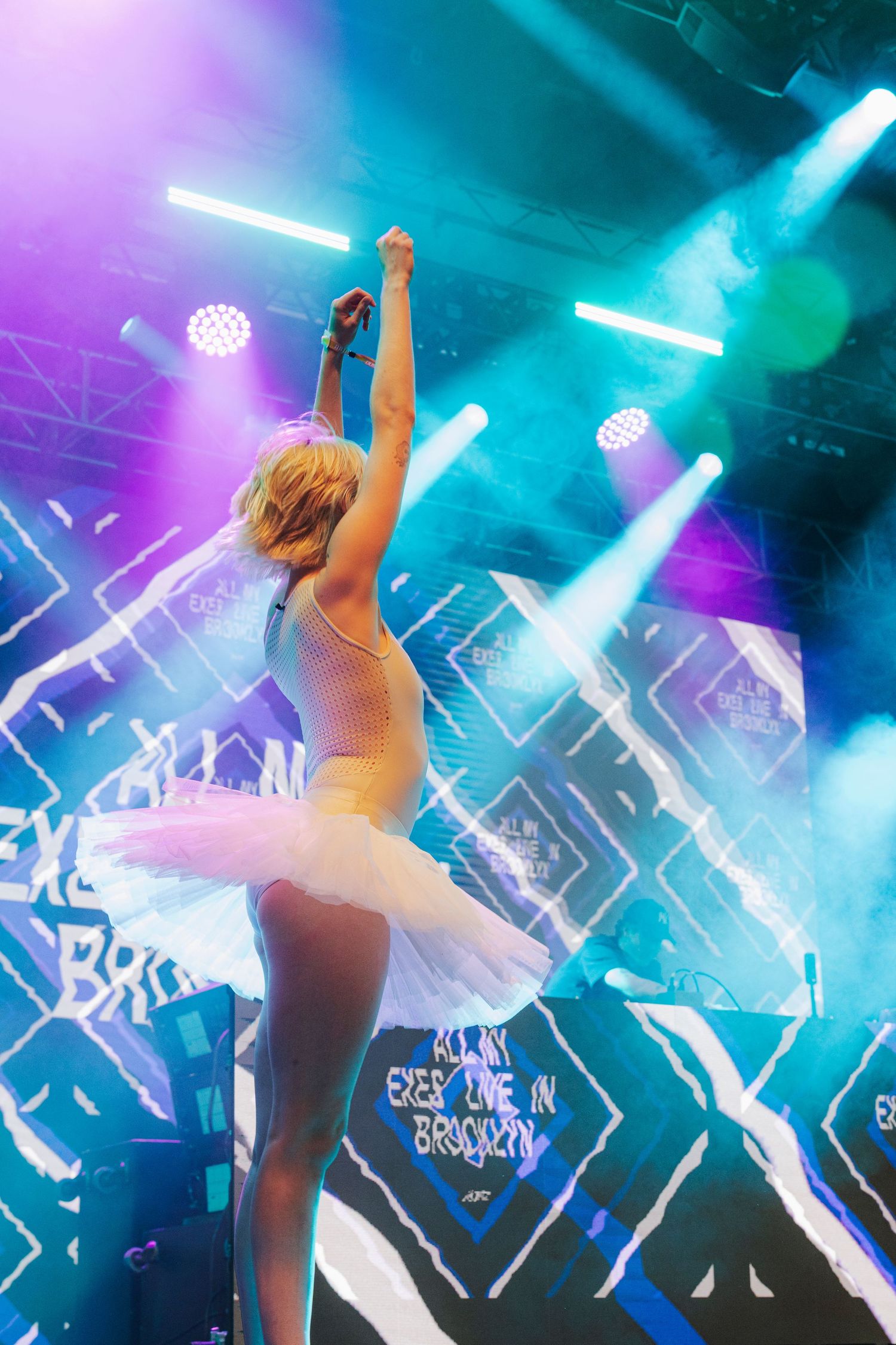Blonde performer in white tutu raising fist on stage with blue neon lights at Brooklyn concert.