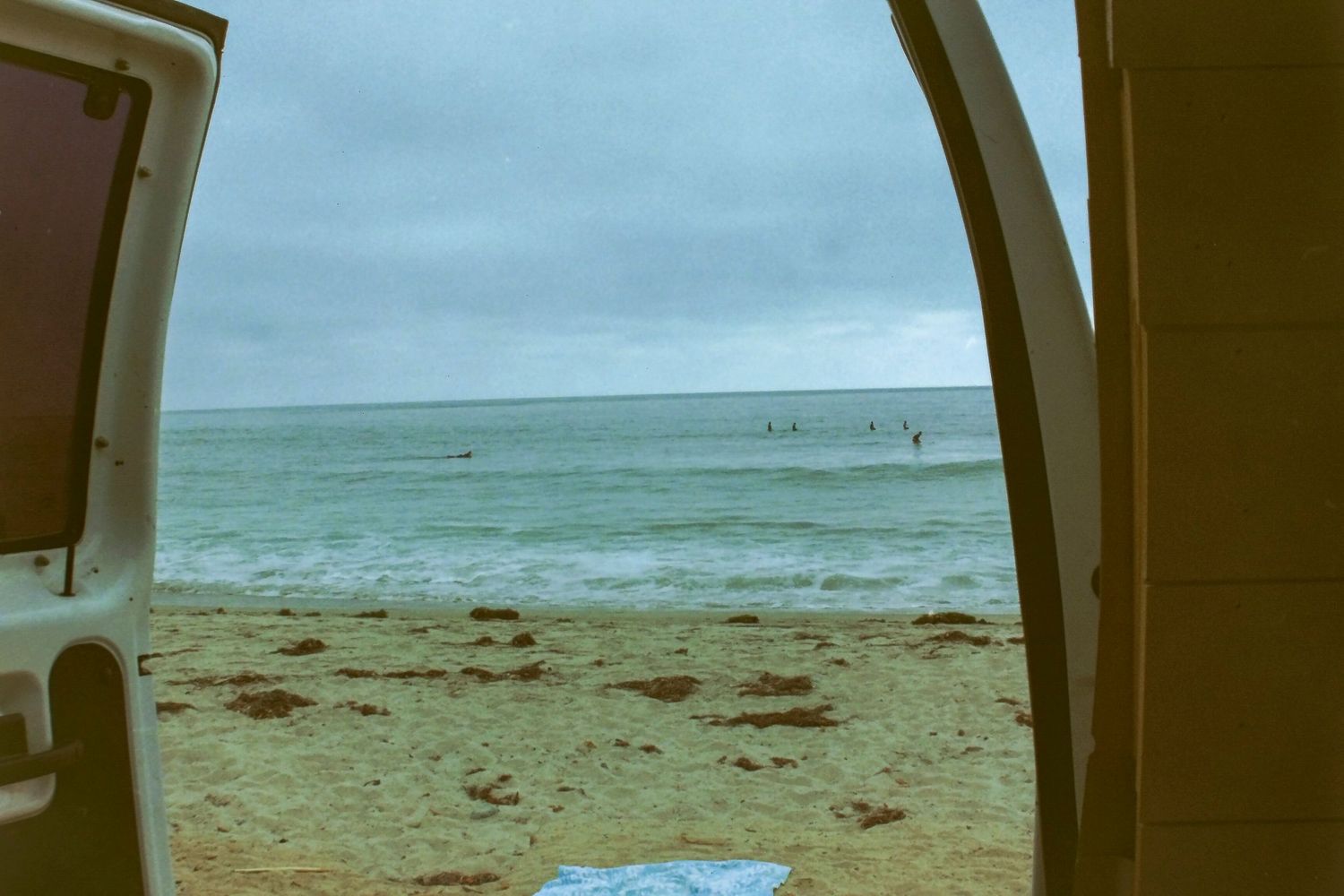 Ocean beach view through open van doors with surfers in the water under a cloudy sky.