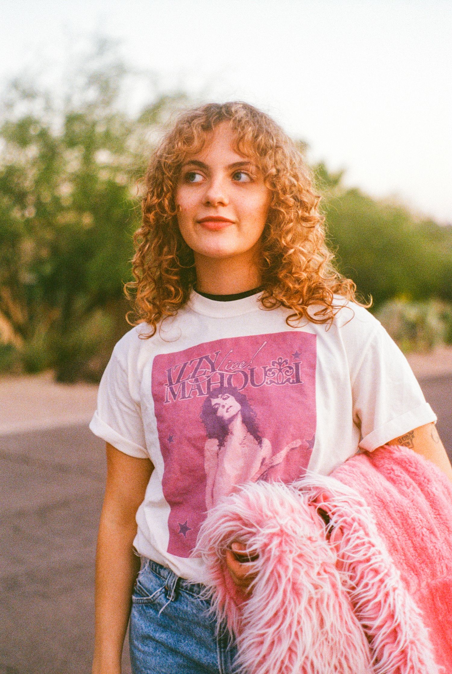 Young woman with curly auburn hair holds pink fluffy jacket wearing a graphic tee outdoors.