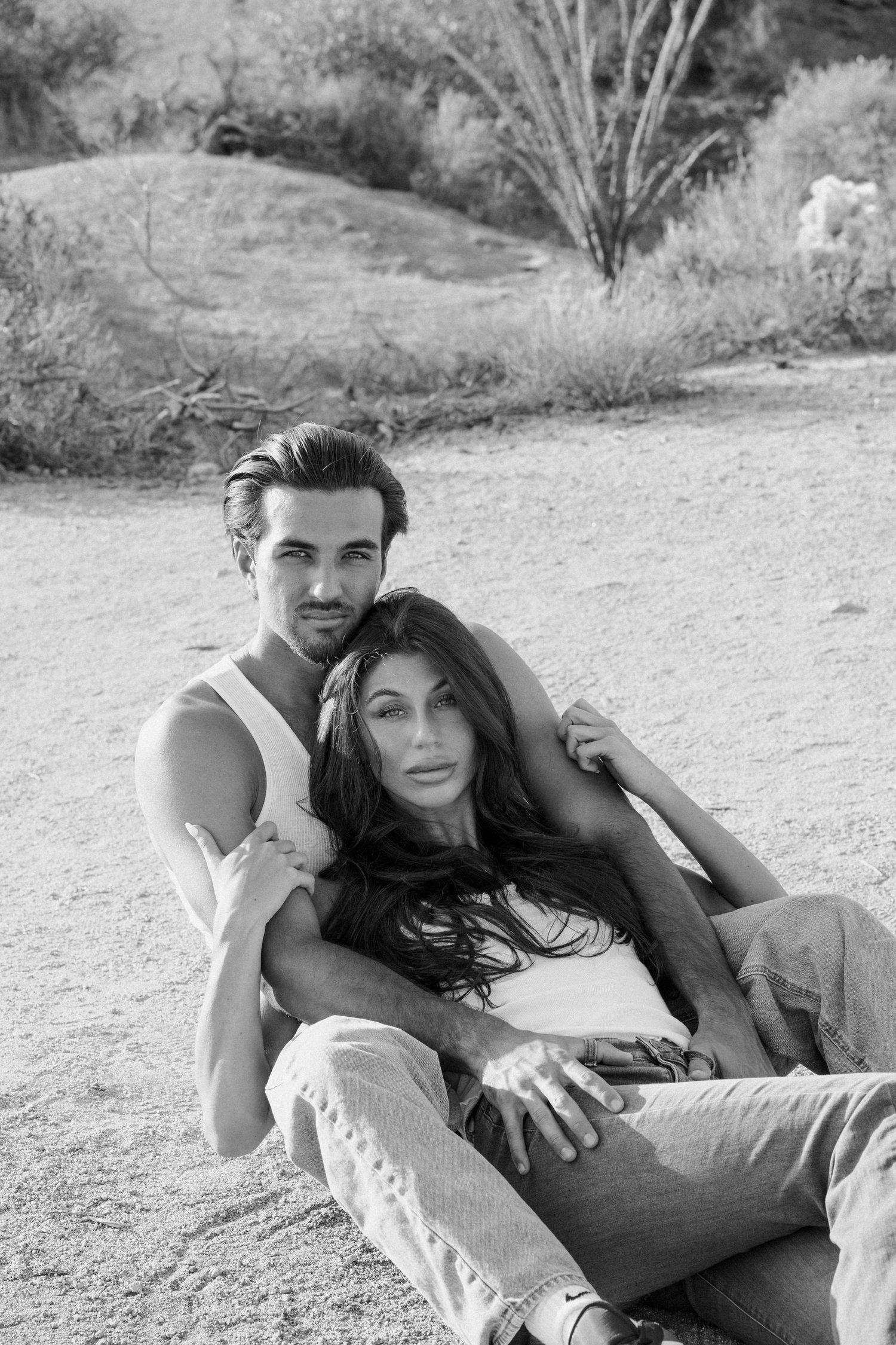 Black and white photo of a couple sitting together outdoors in a desert landscape, man embracing woman from behind.