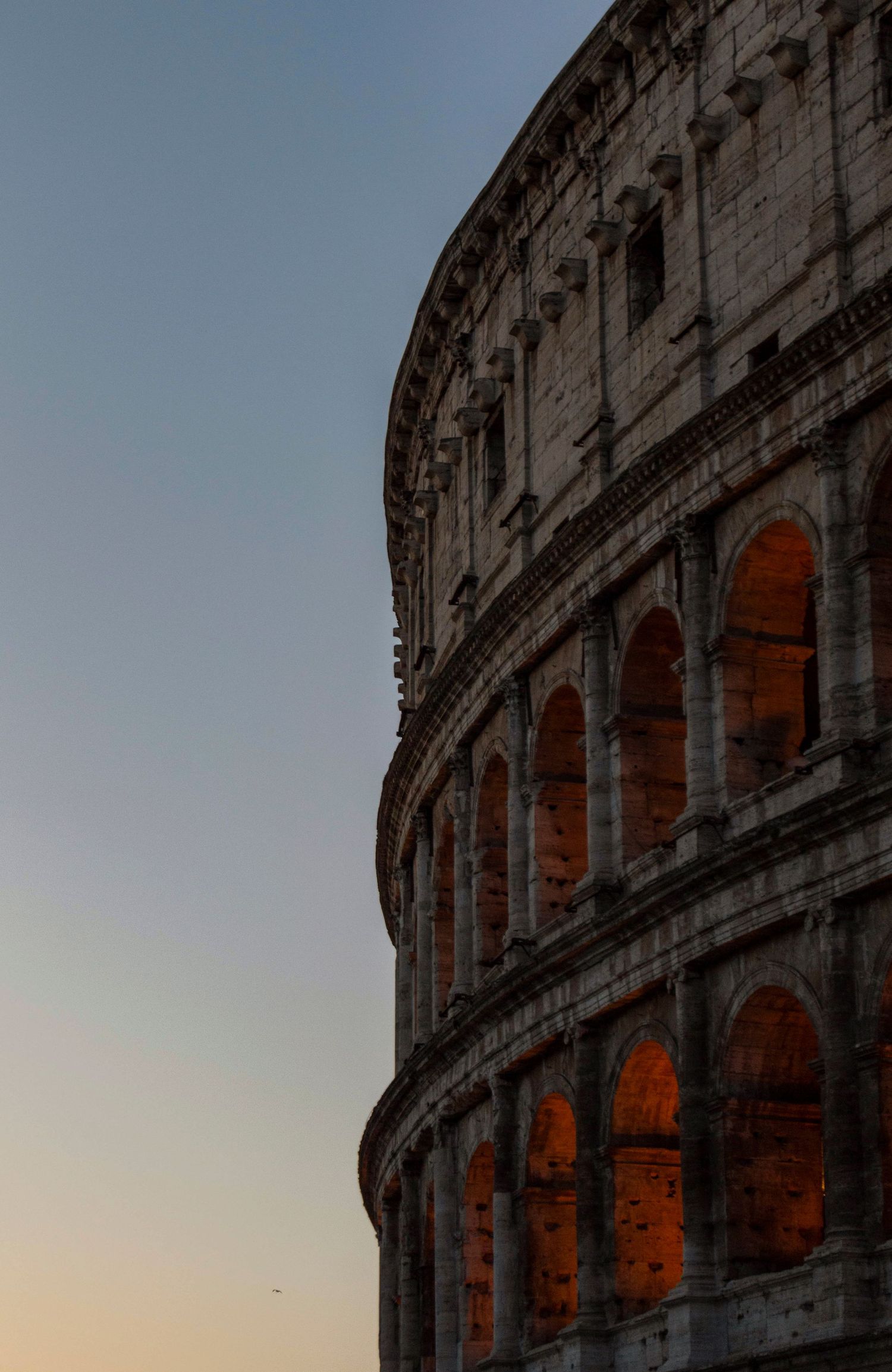 The ancient Roman Colosseum illuminated in warm golden light at dusk against a clear twilight sky.