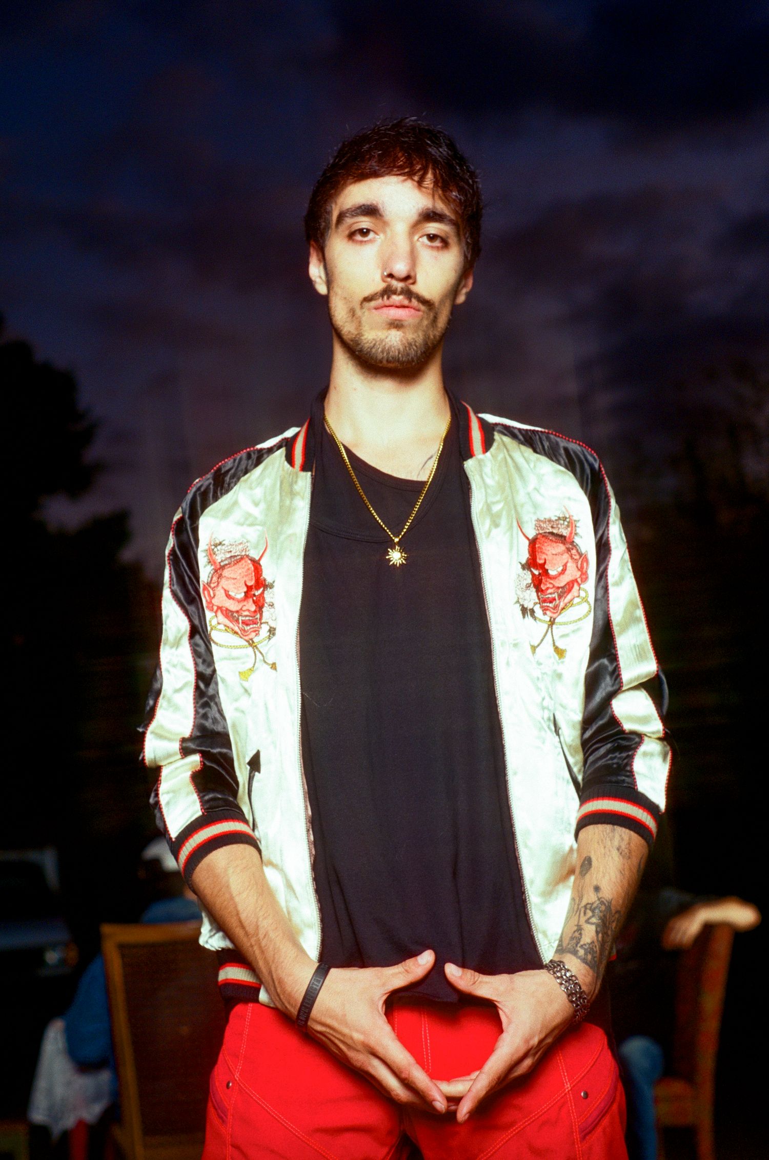 Young man in floral embroidered satin bomber jacket and red pants poses outdoors at night.