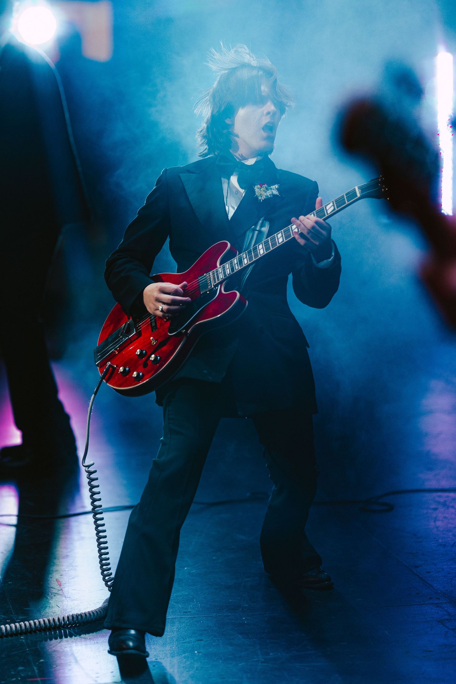 Male guitarist in a black suit playing a red semi-hollow guitar on a smoky blue-lit stage.