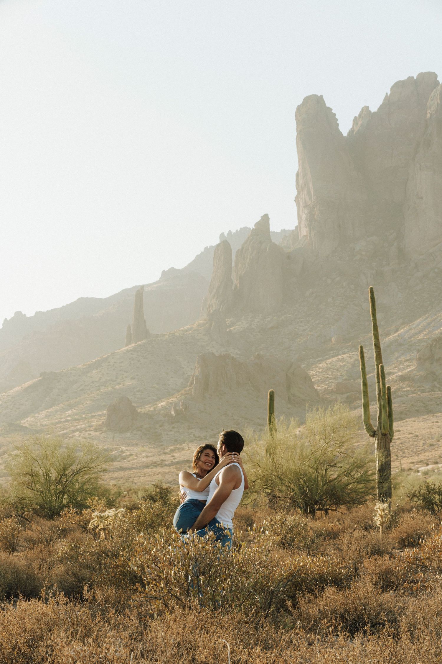 Couple embracing in Arizona desert landscape with saguaro cactus and dramatic rocky mountains at golden hour.