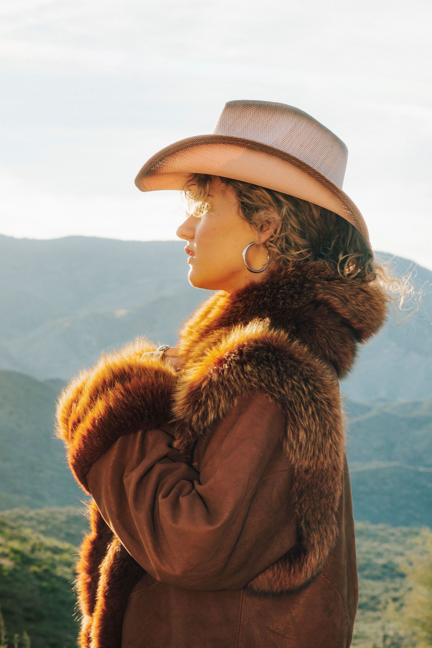 Woman in cowboy hat and fur-trimmed coat poses against mountain landscape in golden sunset light.