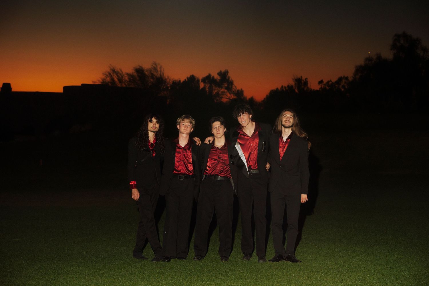 Five people in matching red and black outfits pose together on a grass field at sunset. Band: Garrett James and the Wanderers